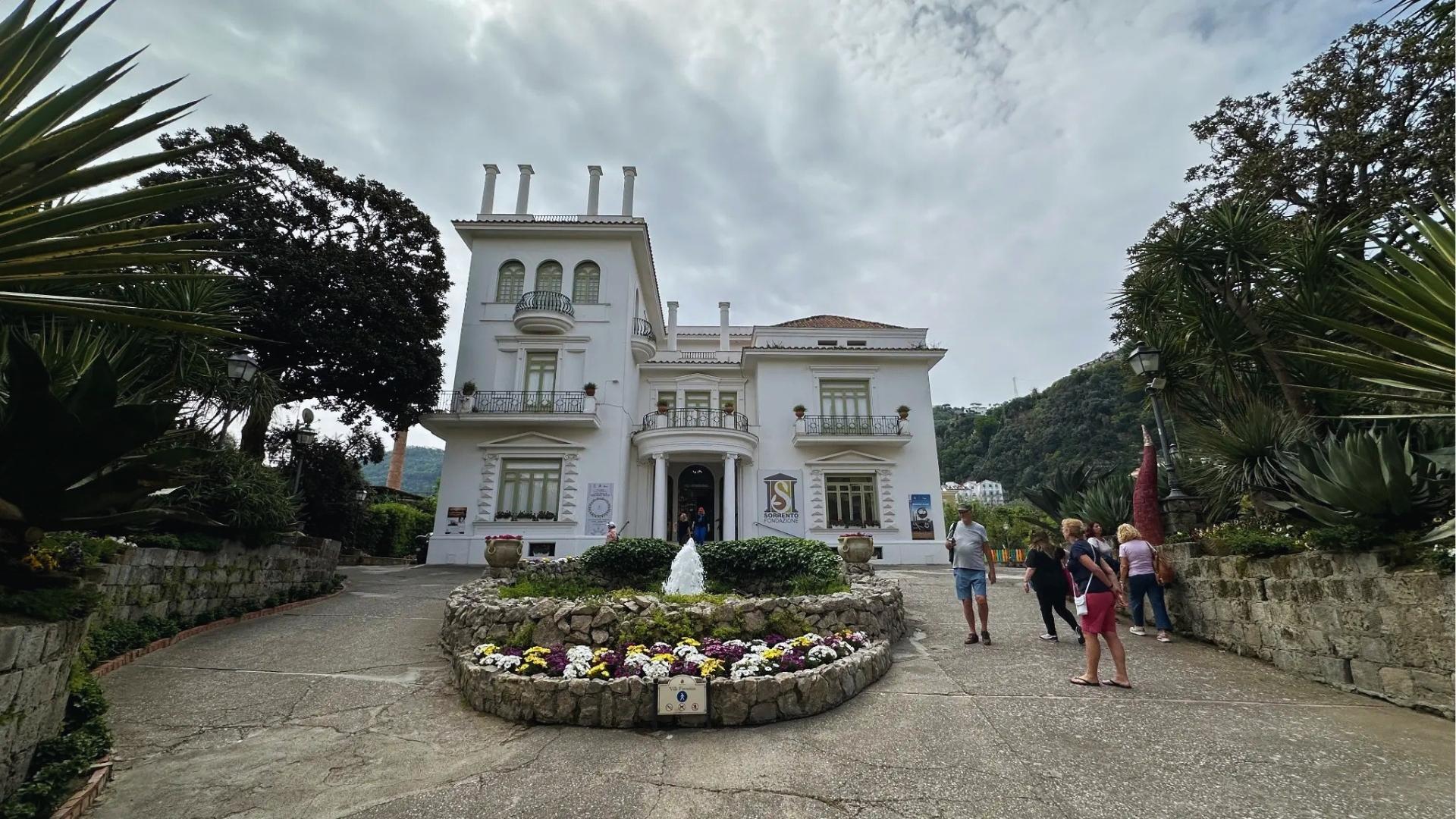 A white mansion with a decorative fountain in front, surrounded by lush greenery.