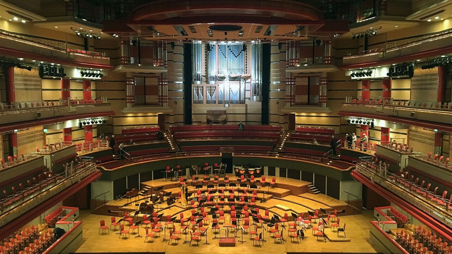 Interior view of a grand auditorium featuring a large orchestra performing, with rows of seats visible in the background.