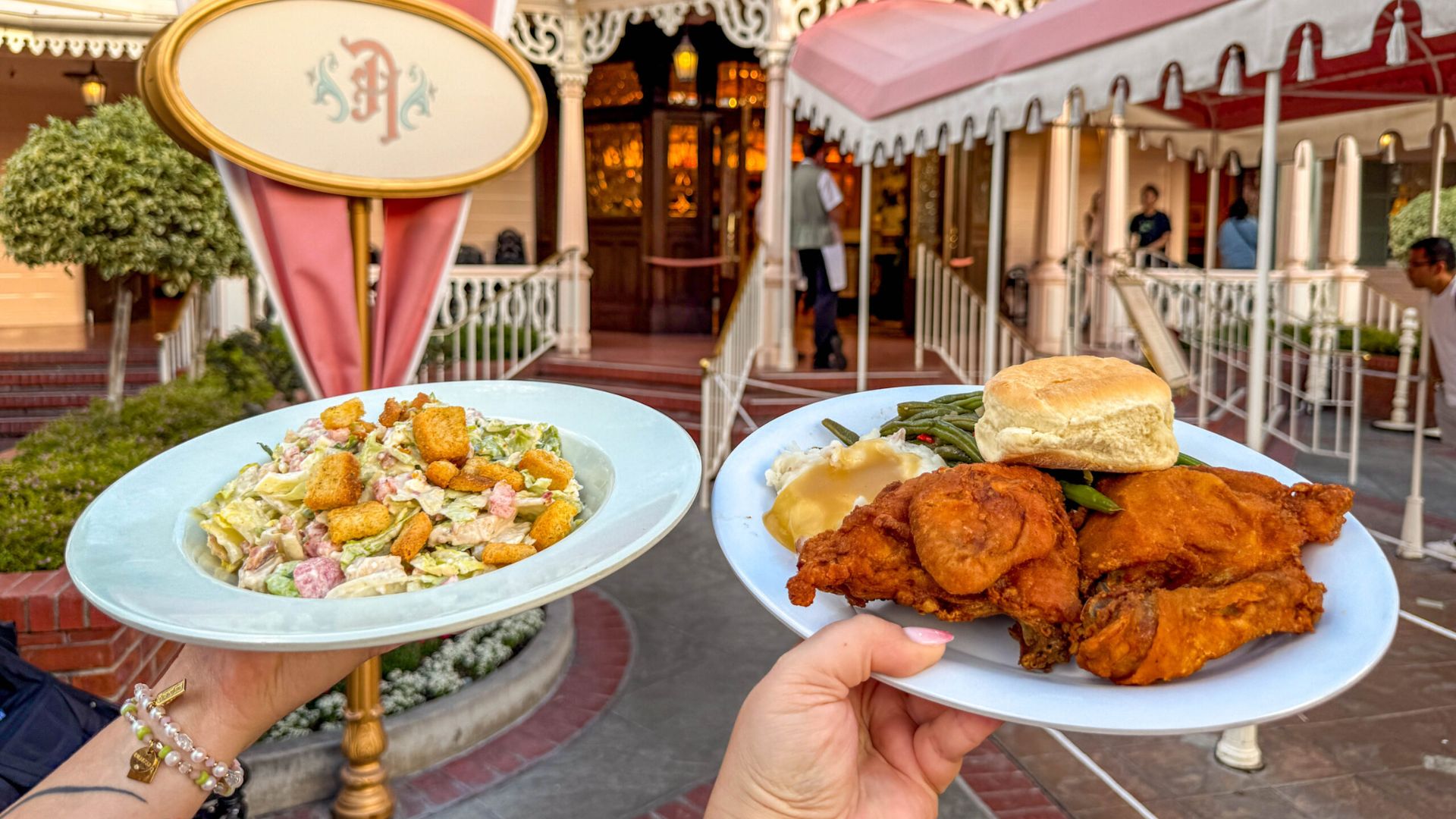 Two people smiling while holding plates of food at Disneyland, surrounded by colorful attractions and festive atmosphere.