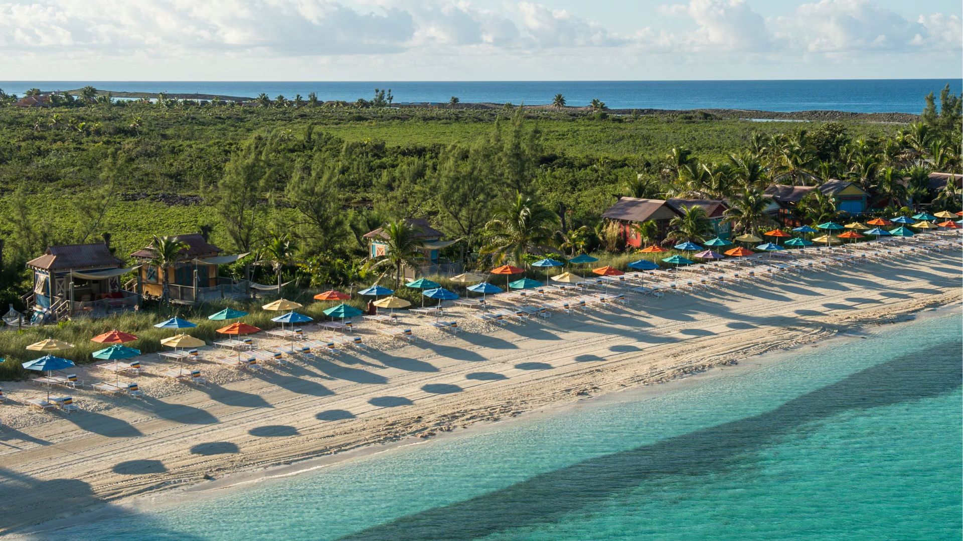 Aerial view of a beach with golden sand and a resort, showcasing clear blue water and sunbathers enjoying the sun.
