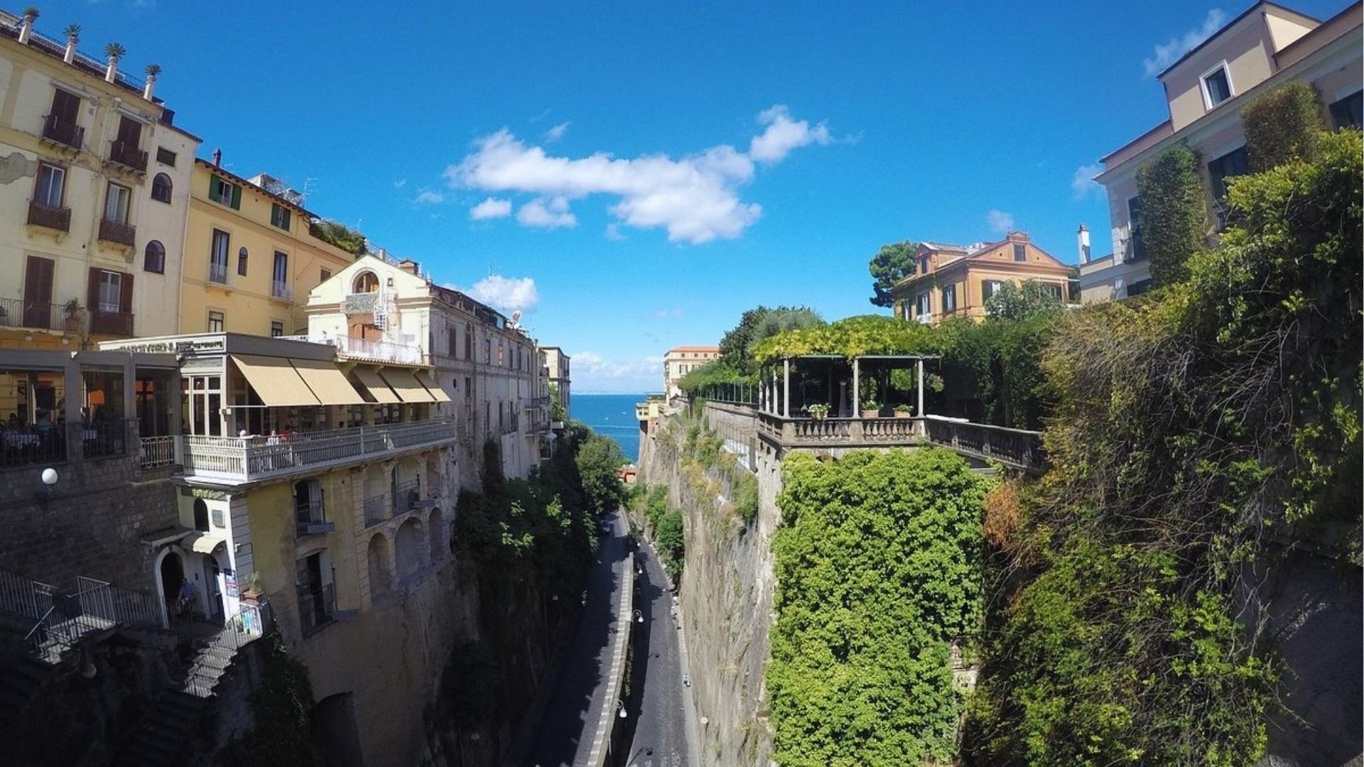 A narrow street lined with buildings, leading to a view of the sea in the background.