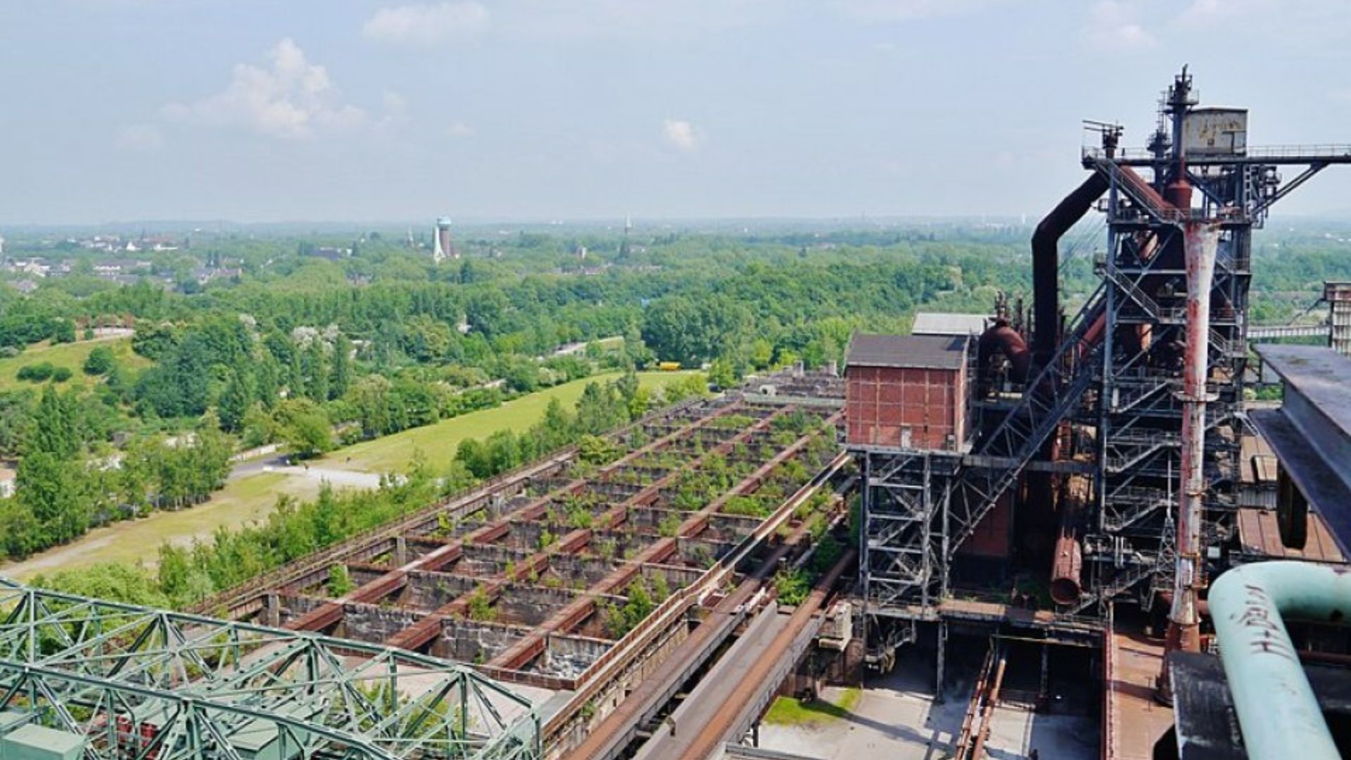 A steel mill stands in the background, surrounded by lush green trees, creating a contrast between industry and nature.