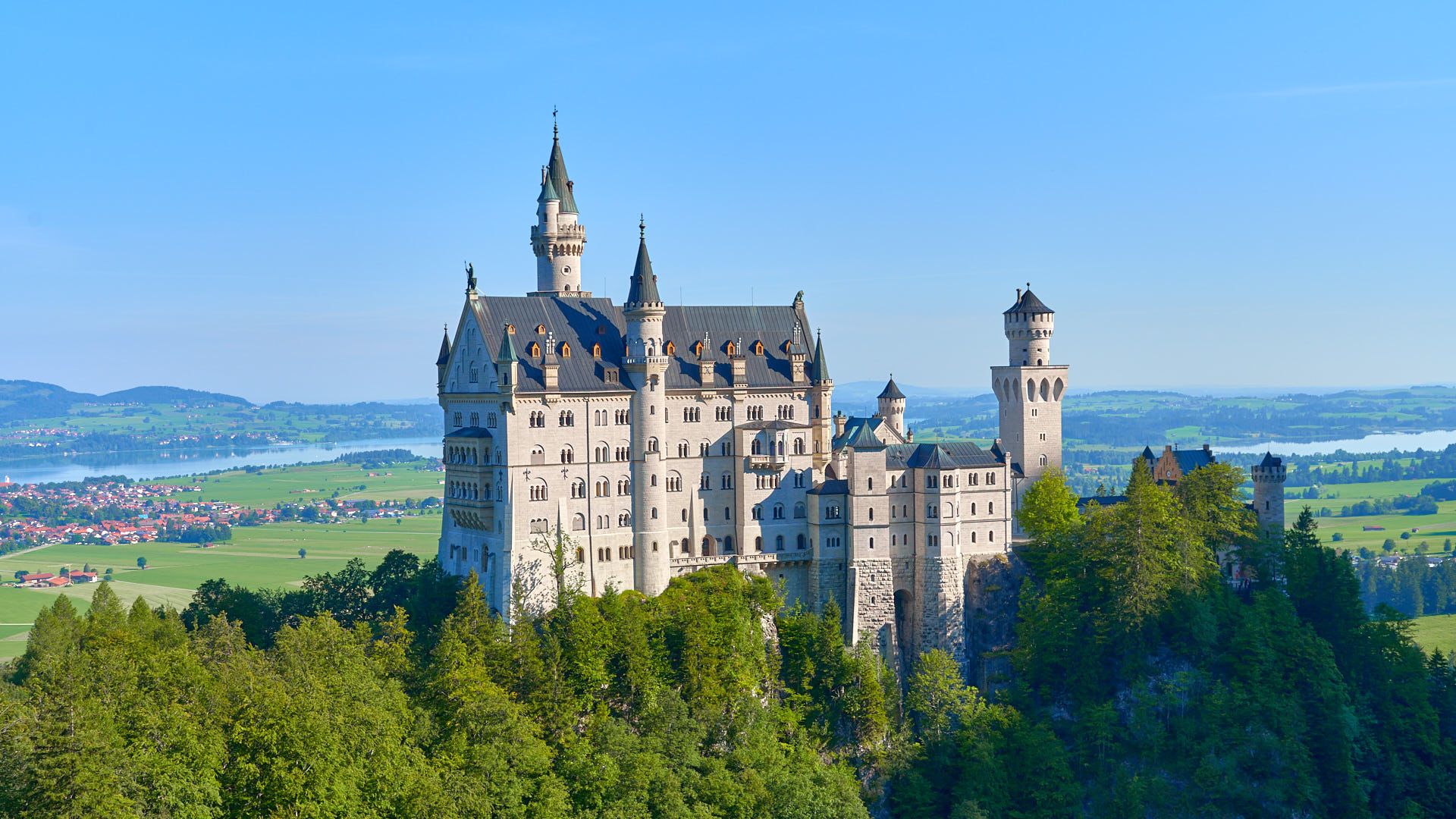 Neuschwanstein Castle in Germany, a fairy-tale structure surrounded by lush forests and mountains under a clear blue sky.