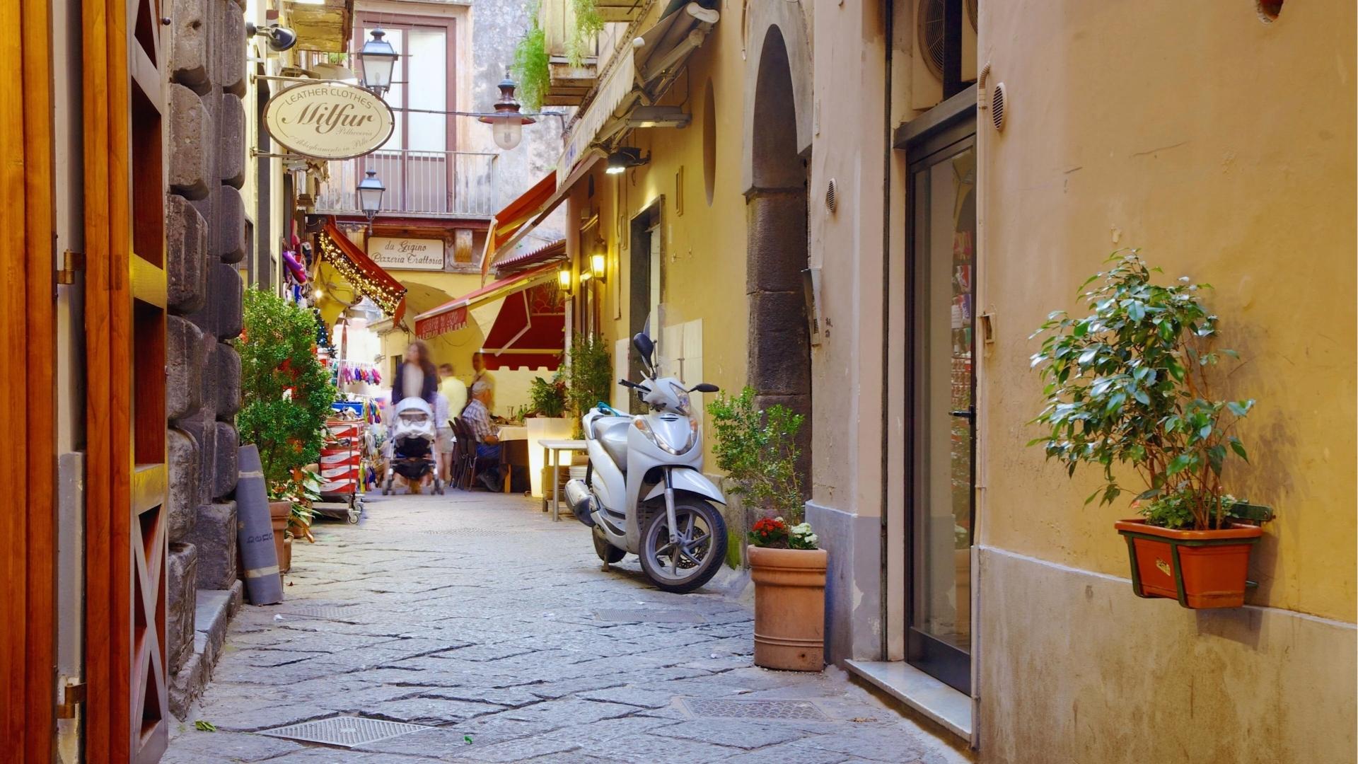 A narrow street featuring a motorcycle parked along the side, surrounded by buildings and a clear blue sky.