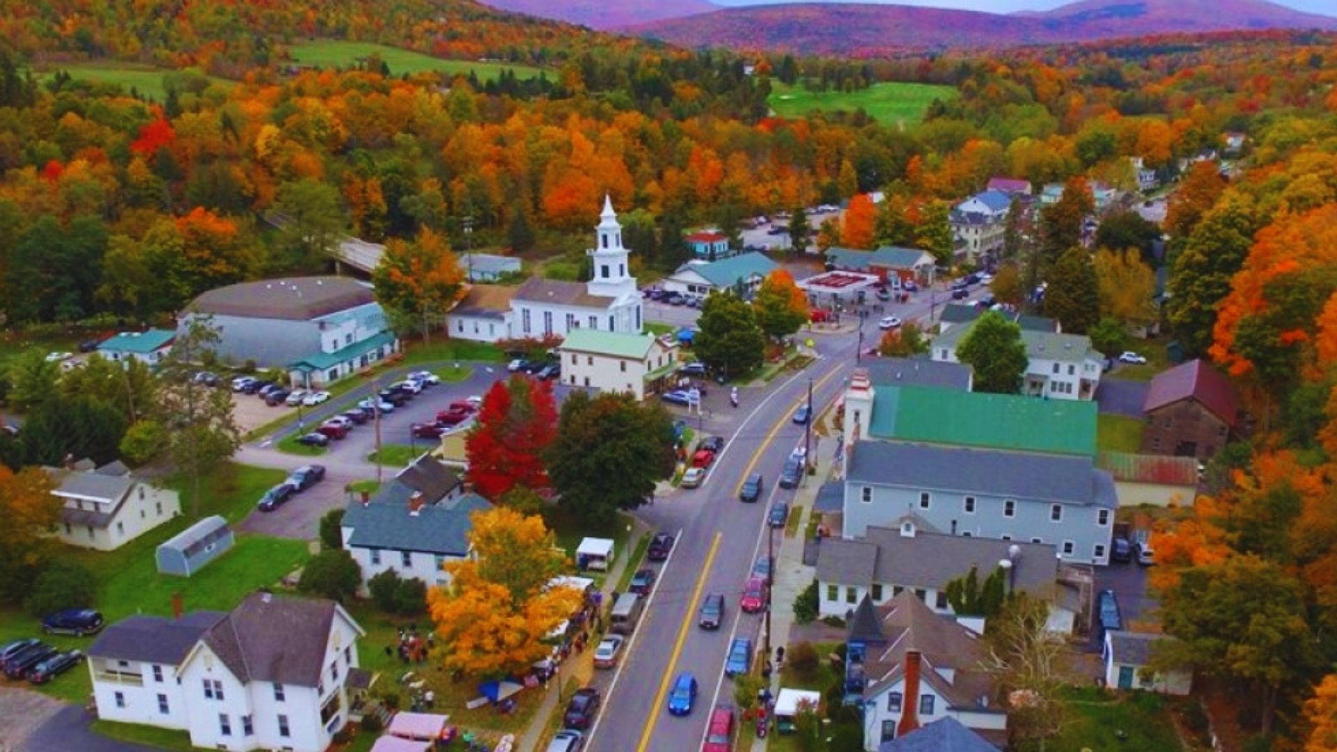 Aerial view of a small town surrounded by vibrant fall foliage, showcasing colorful trees and quaint buildings.
