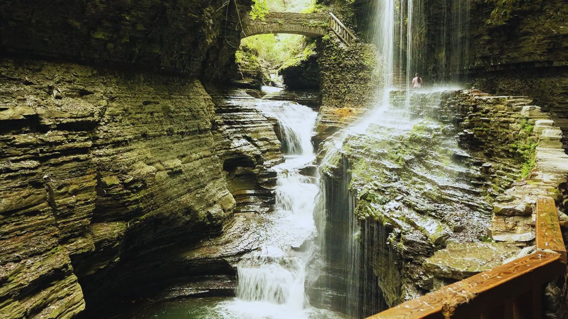 A waterfall cascades down a narrow path, surrounded by lush greenery and rocks.