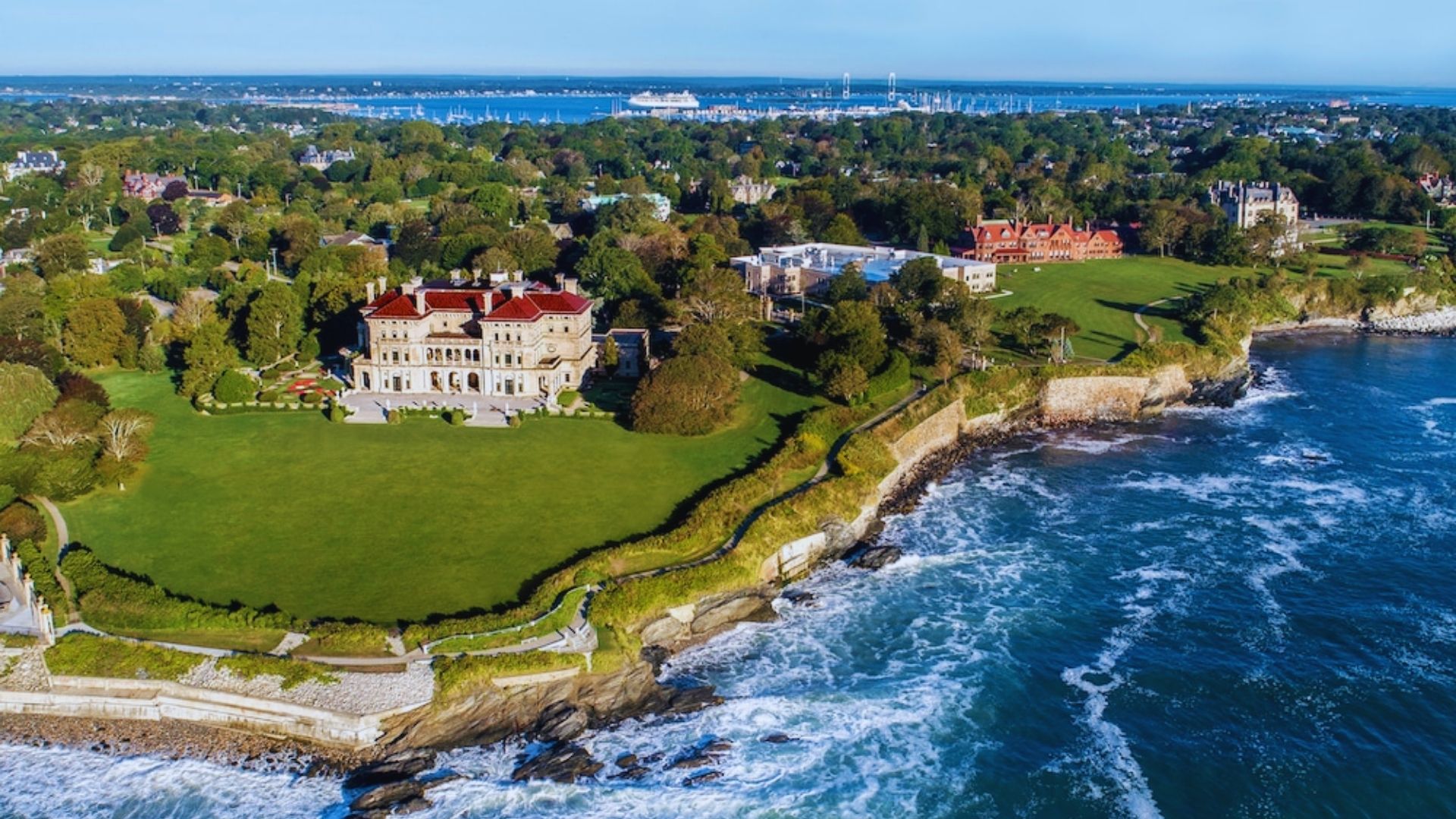 Aerial view of a grand mansion situated along the shoreline, surrounded by lush greenery and blue waters.