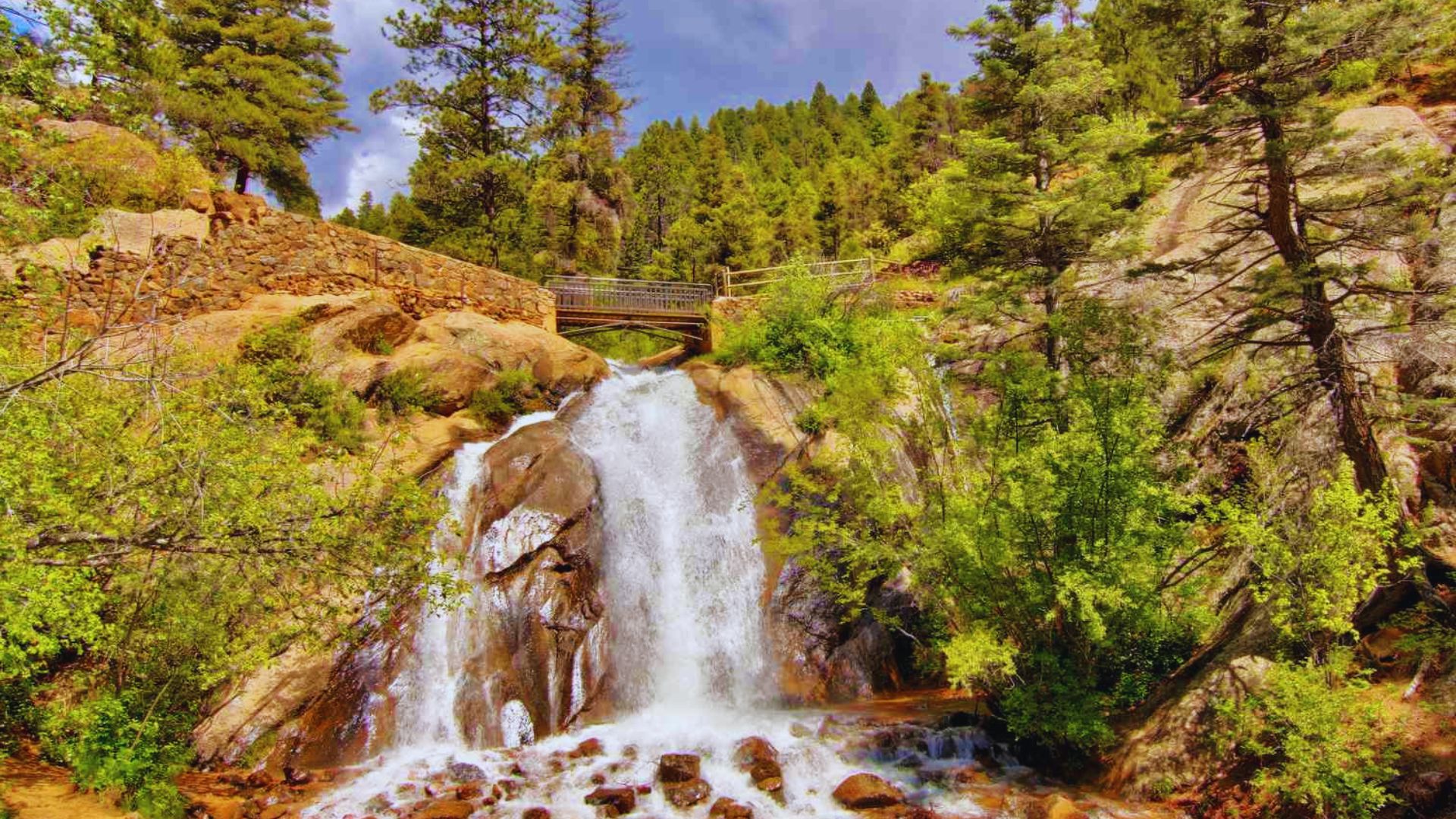 A scenic waterfall cascades down rocky cliffs, surrounded by lush green trees in a mountainous landscape.