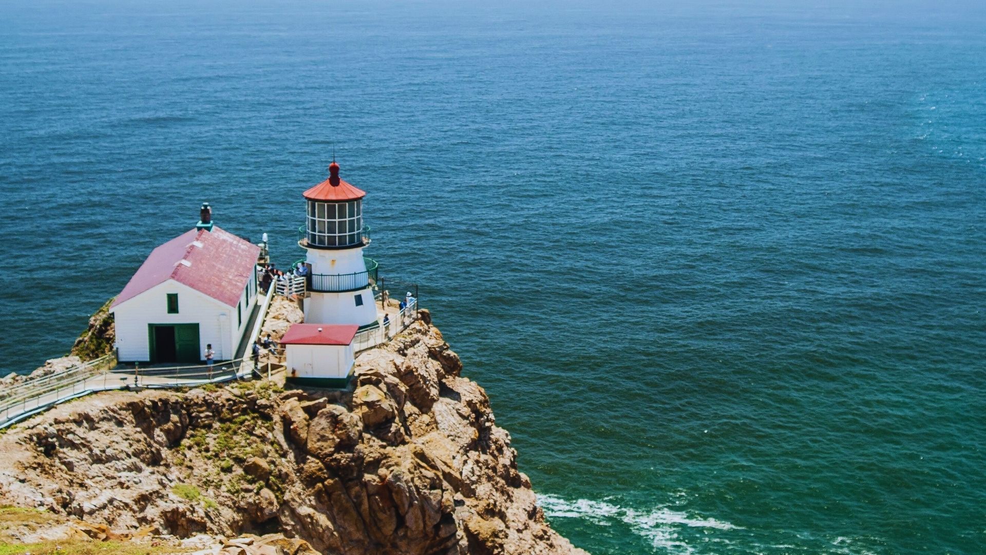 A lighthouse stands on a cliff, overlooking a vast ocean under a clear blue sky.