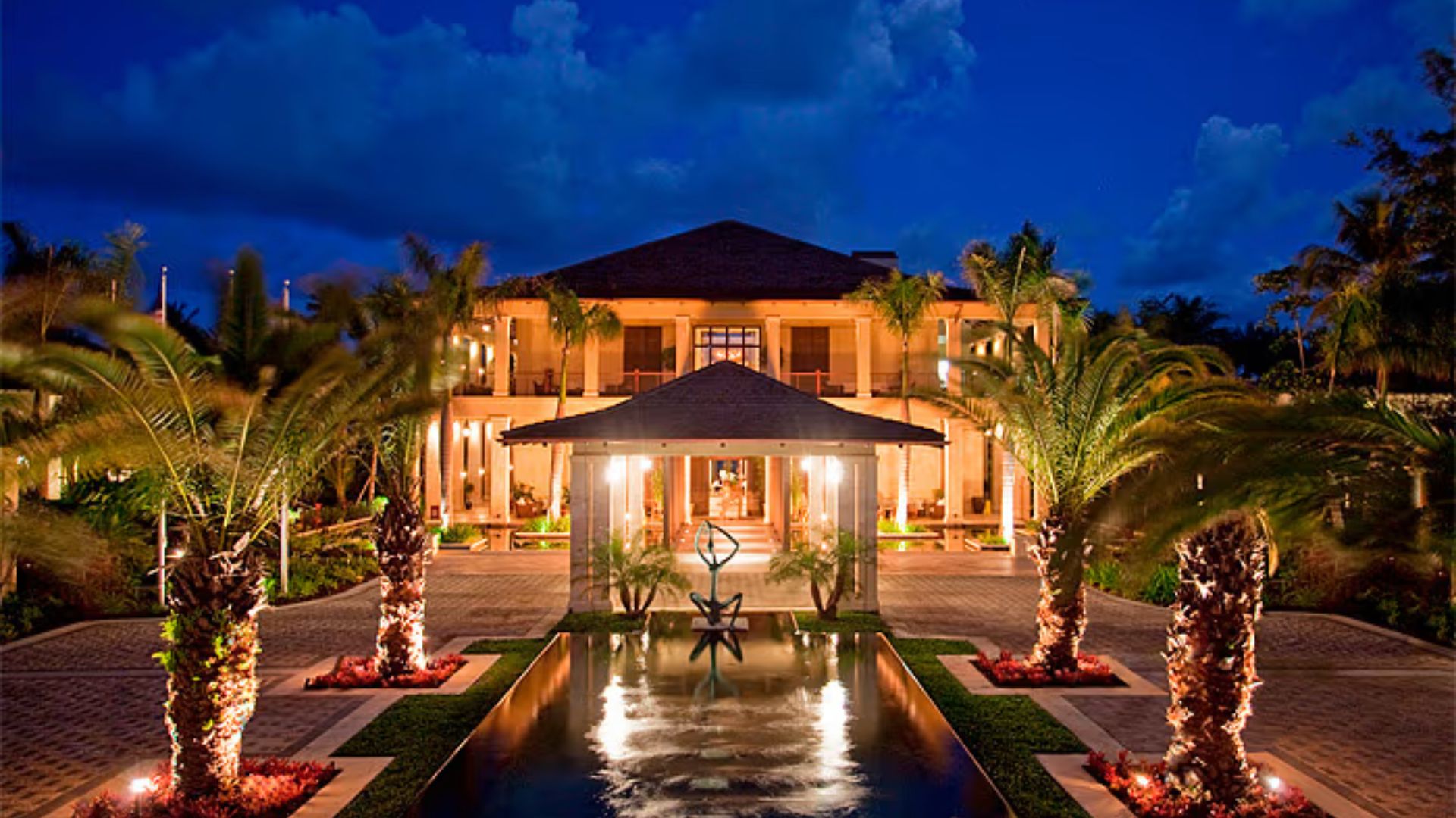 A large home surrounded by palm trees, featuring a decorative fountain in the front yard.