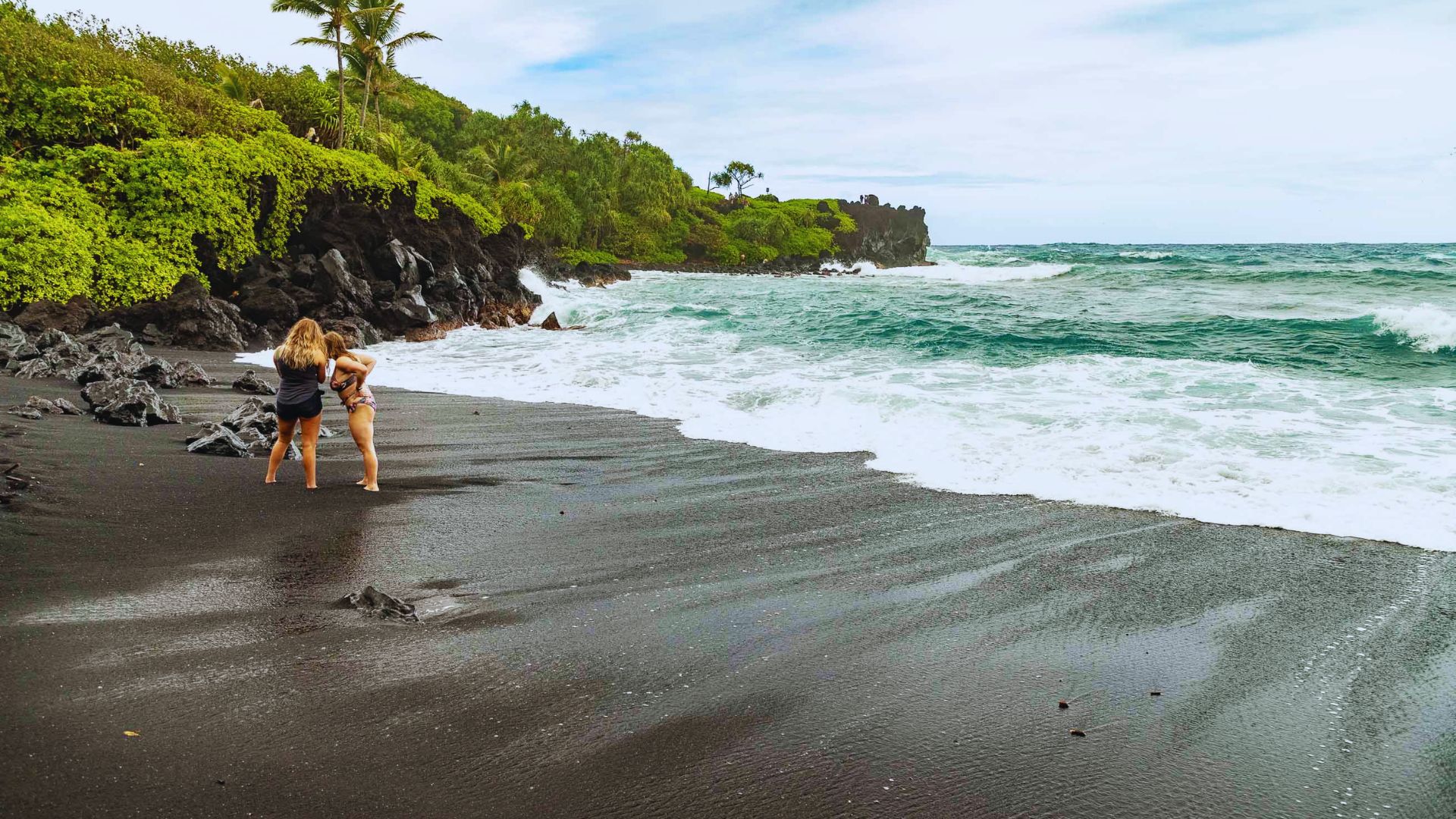 A woman stands on a black sand beach, holding a surfboard, with waves gently crashing in the background.