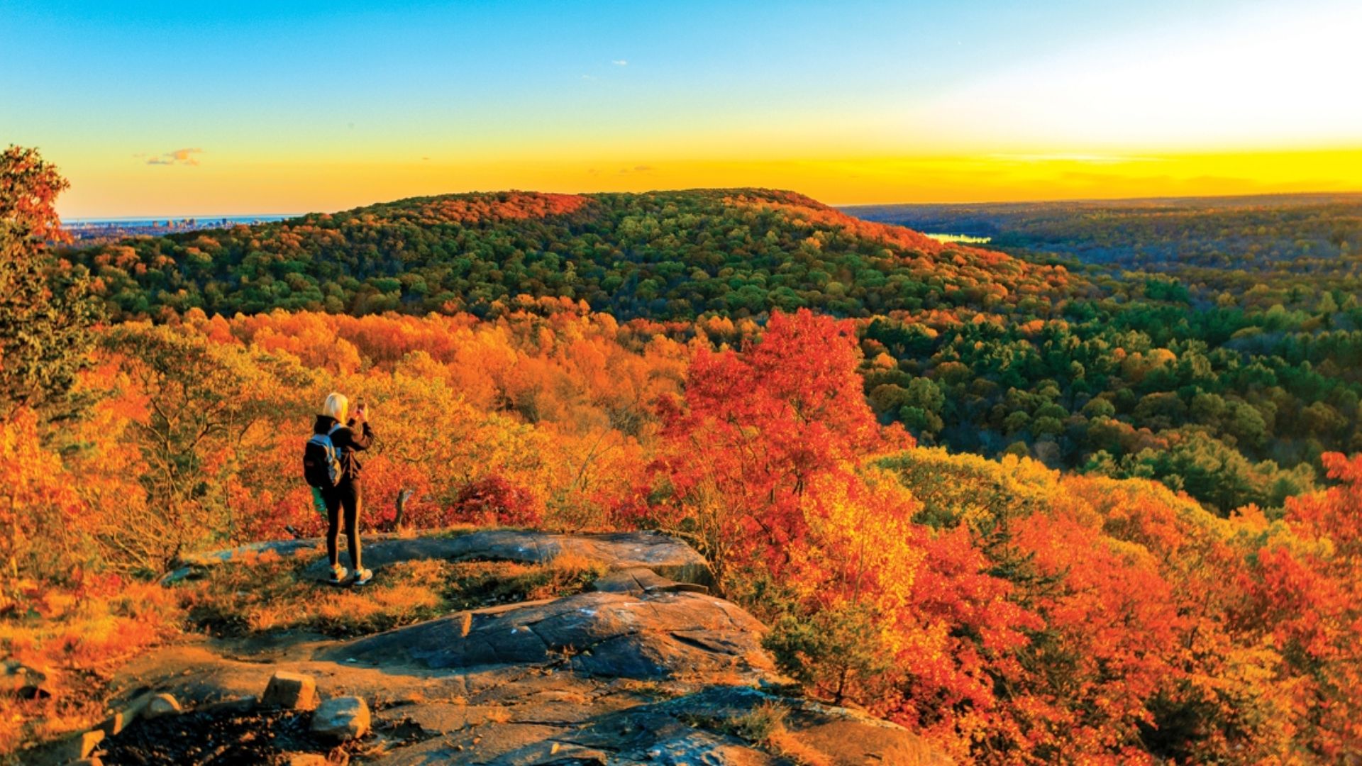 A person stands on a rock, gazing at vibrant fall colors in the surrounding landscape.