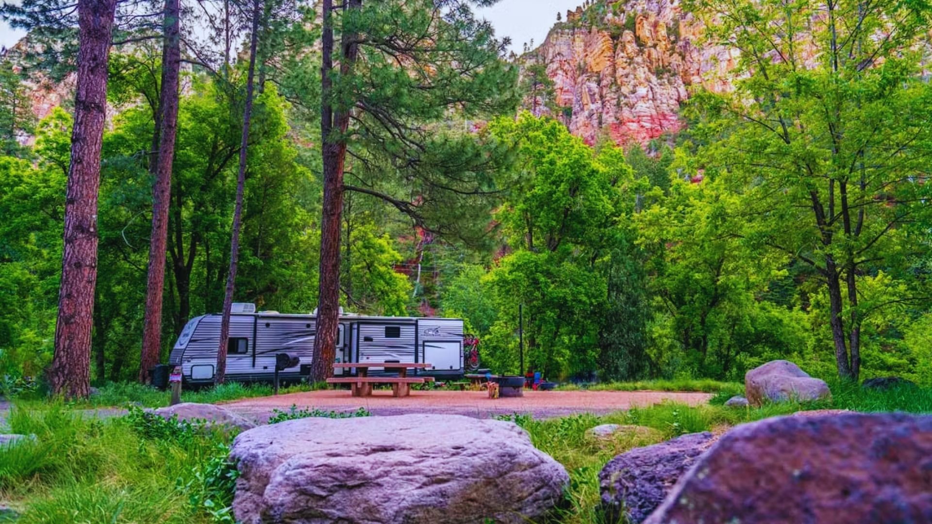 An RV parked in a wooded area beside a picnic table, surrounded by trees and natural scenery.