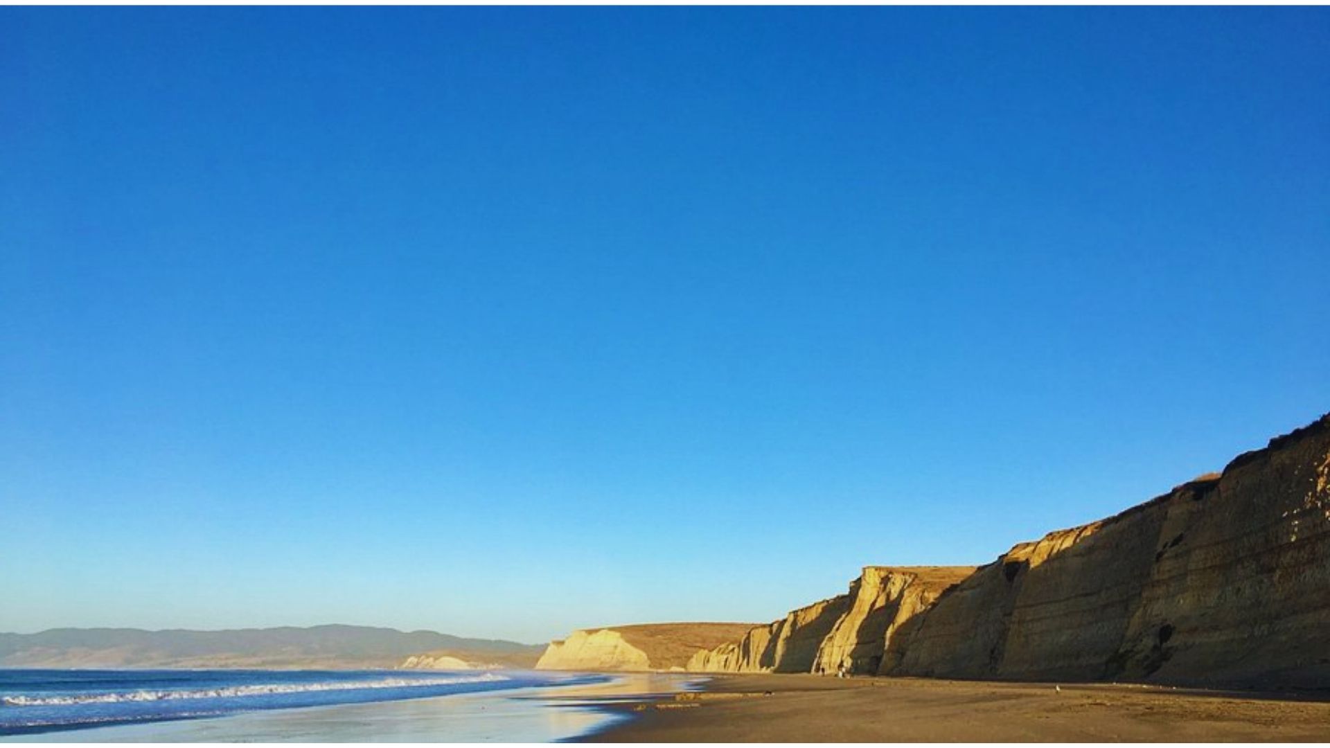 A scenic beach with towering cliffs and gentle waves lapping at the shore under a clear blue sky.
