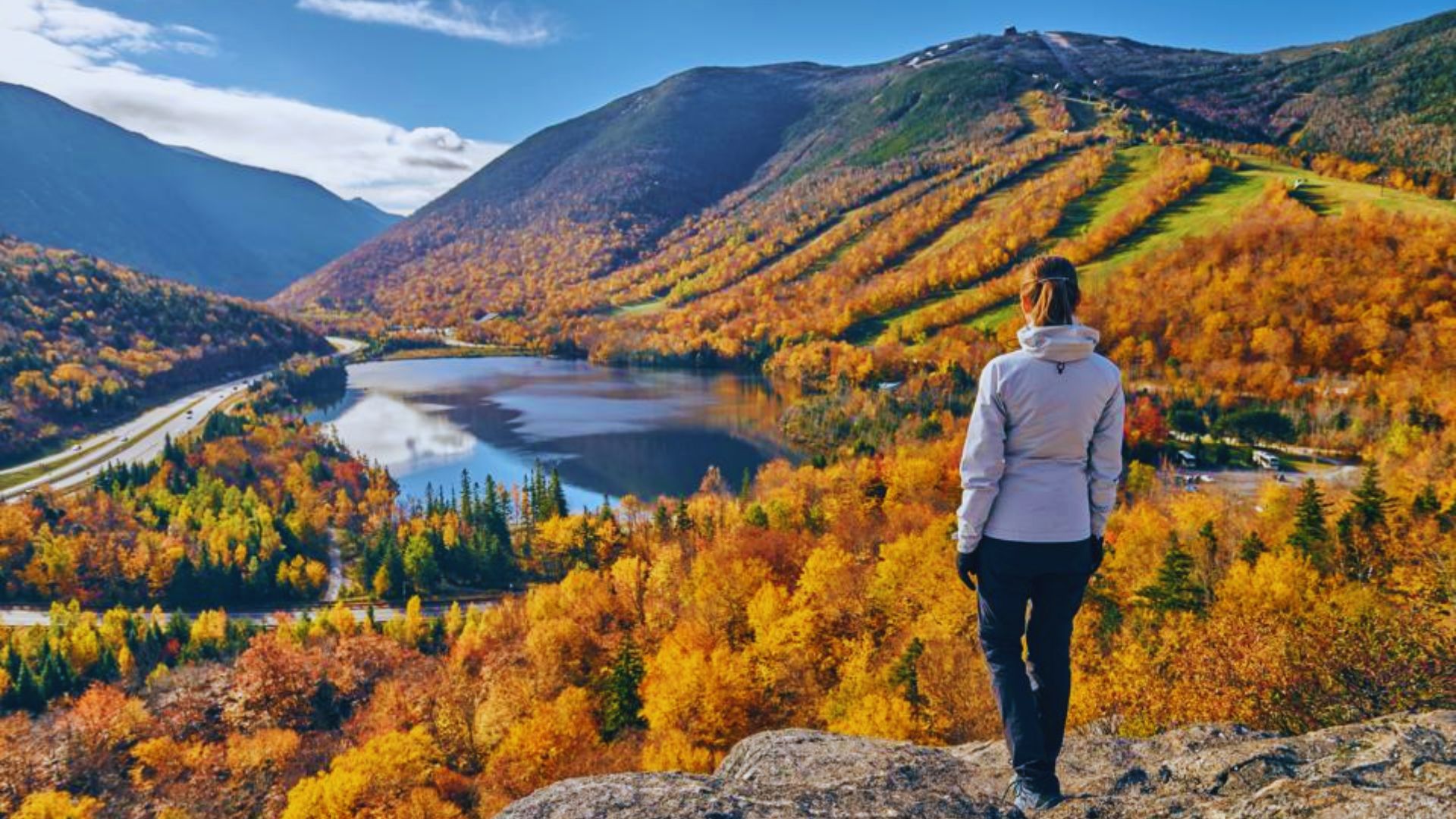 A woman on a mountain peak admires a picturesque lake and surrounding mountains in the distance.