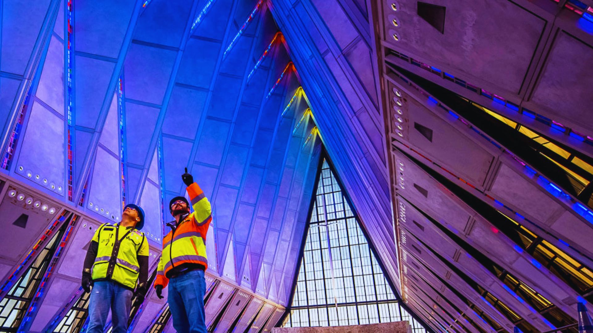 Two men in bright safety vests stand outside a large building, appearing to review plans or discuss work.
