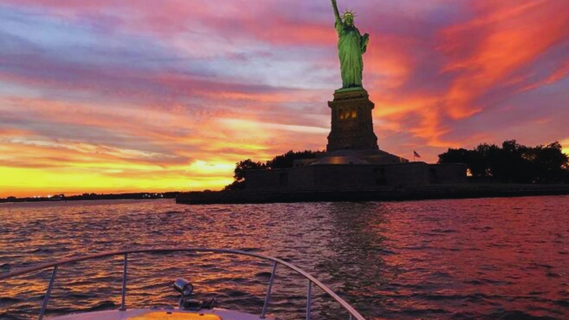 The Statue of Liberty silhouetted against a vibrant sunset, viewed from a boat on the water.