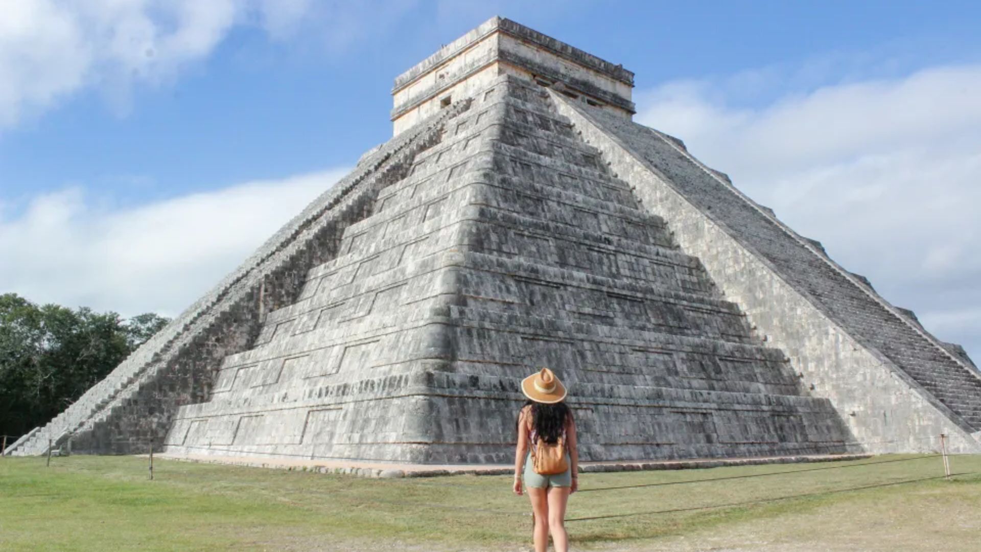 A woman wearing a hat stands in front of a large pyramid, showcasing her interest in the historical landmark.