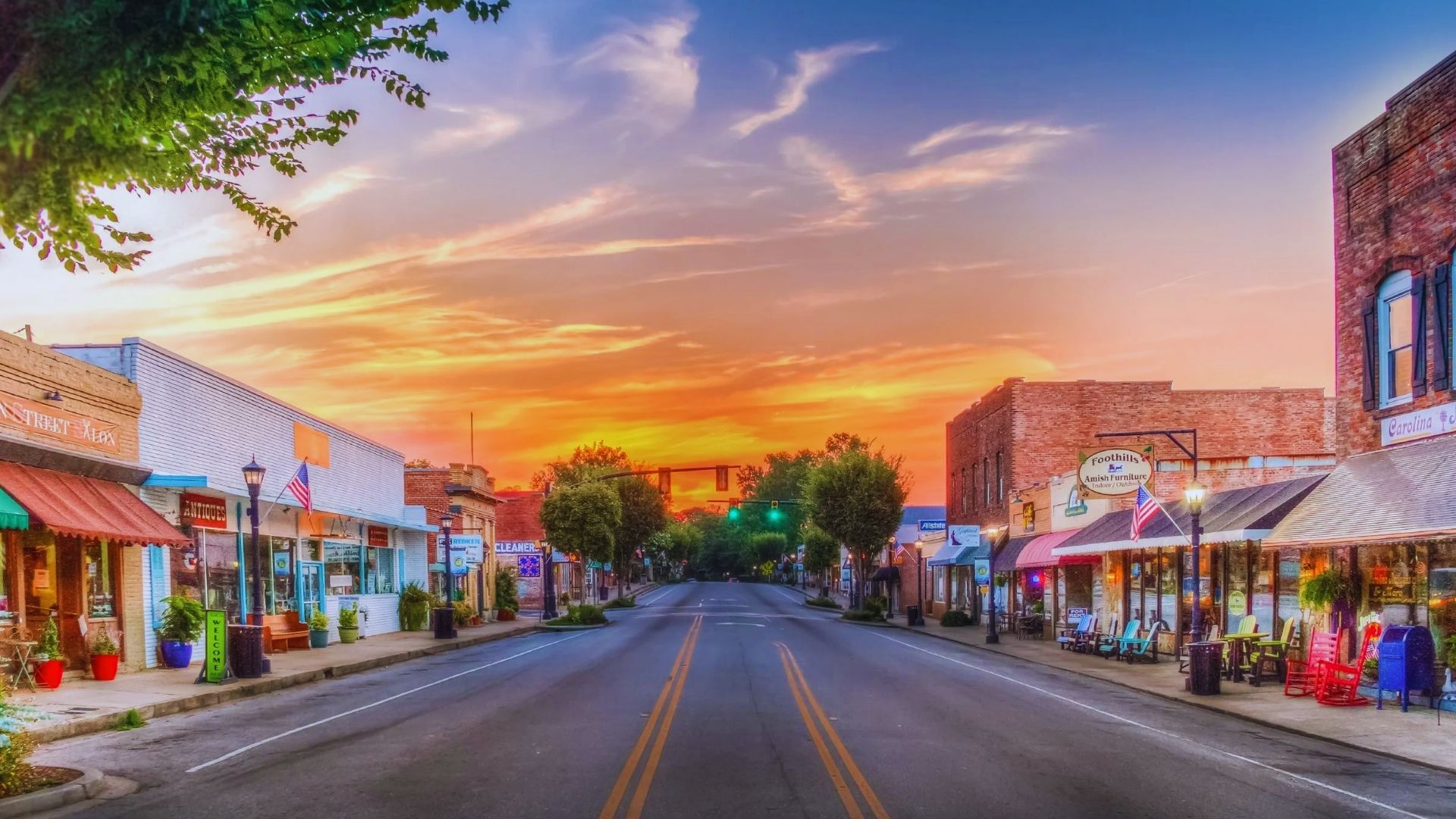 A small town street bathed in warm hues of sunset, with silhouettes of buildings and trees lining the road.