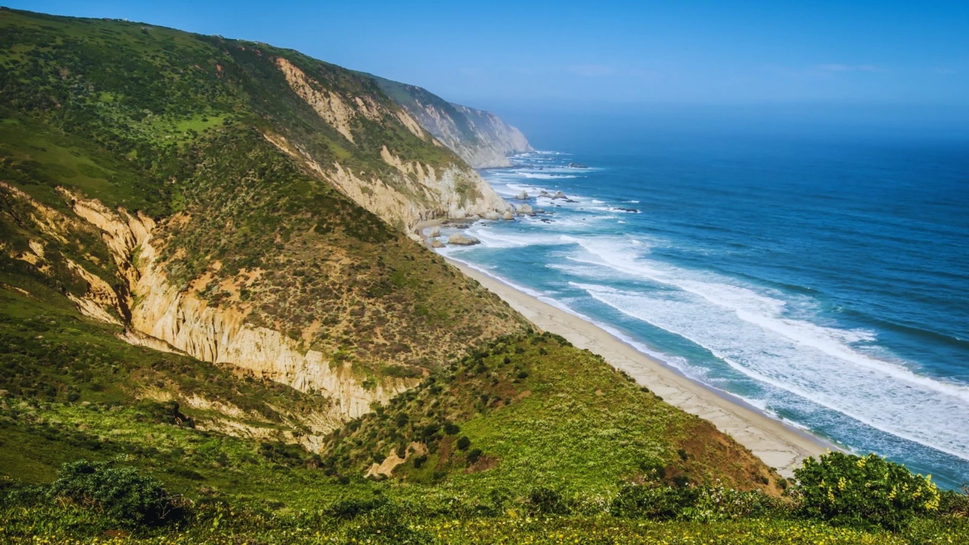 A panoramic view of the Pacific Ocean coast featuring sandy beaches and gentle waves under a bright sunny sky.