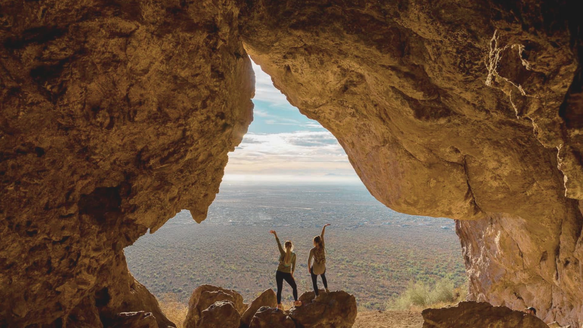 Two people stand in a cave, arms raised, as they celebrate the natural beauty surrounding them.