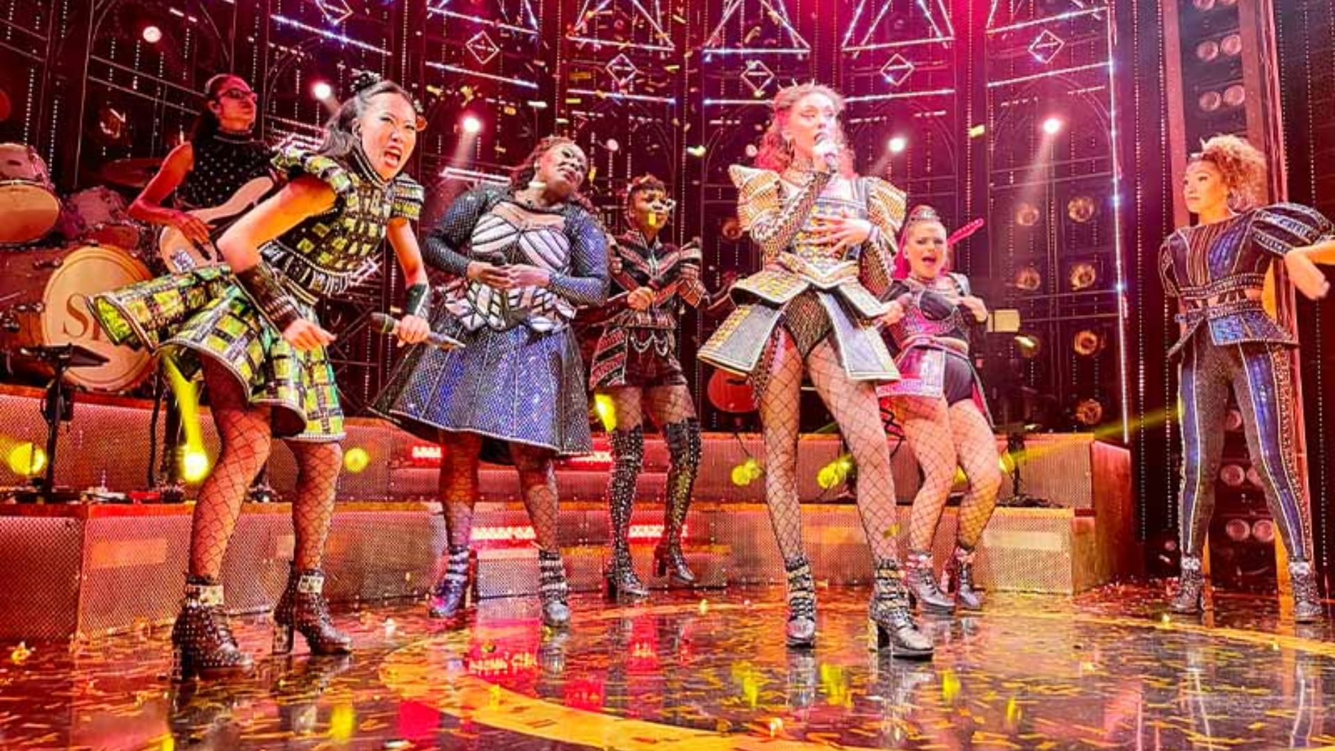 The cast of the musical performs energetically on stage, showcasing vibrant costumes and engaging choreography.