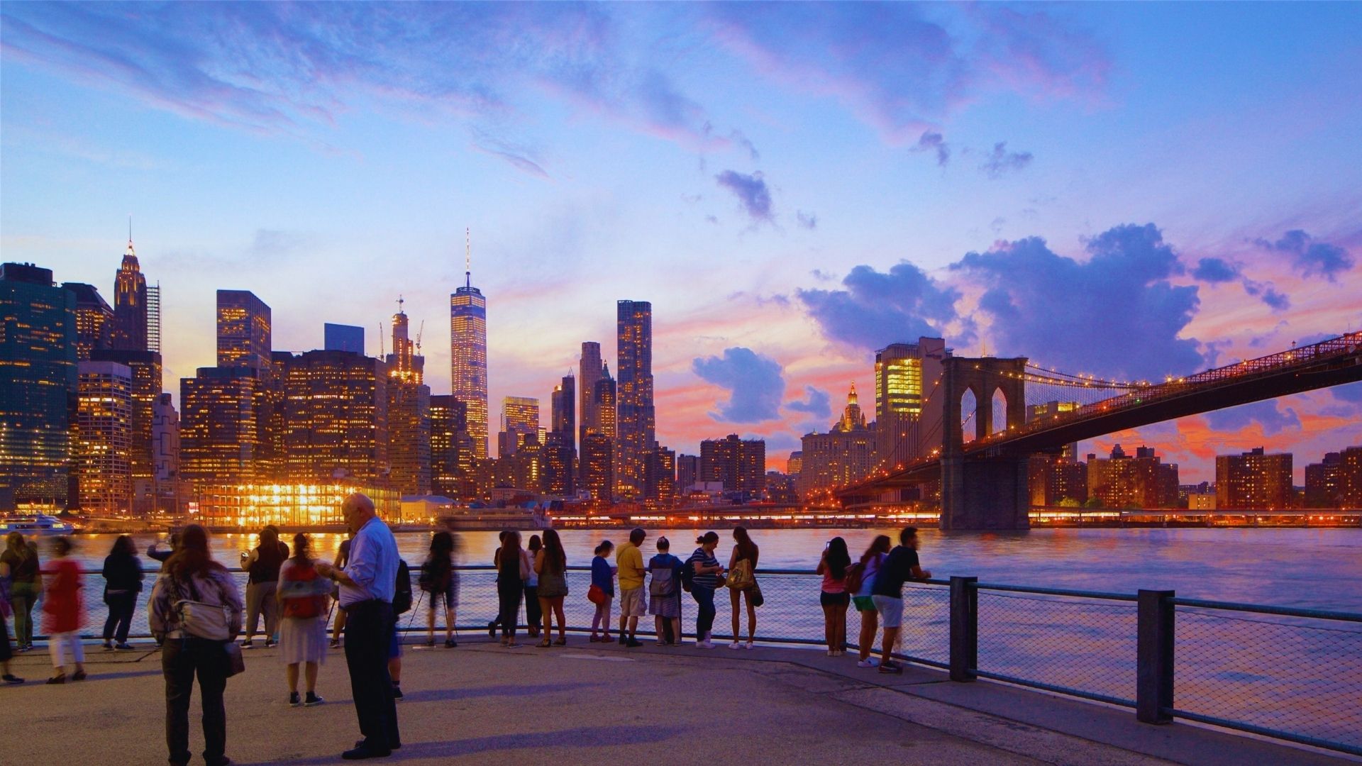 People stand on the waterfront at dusk, silhouetted against a city skyline illuminated by twilight.