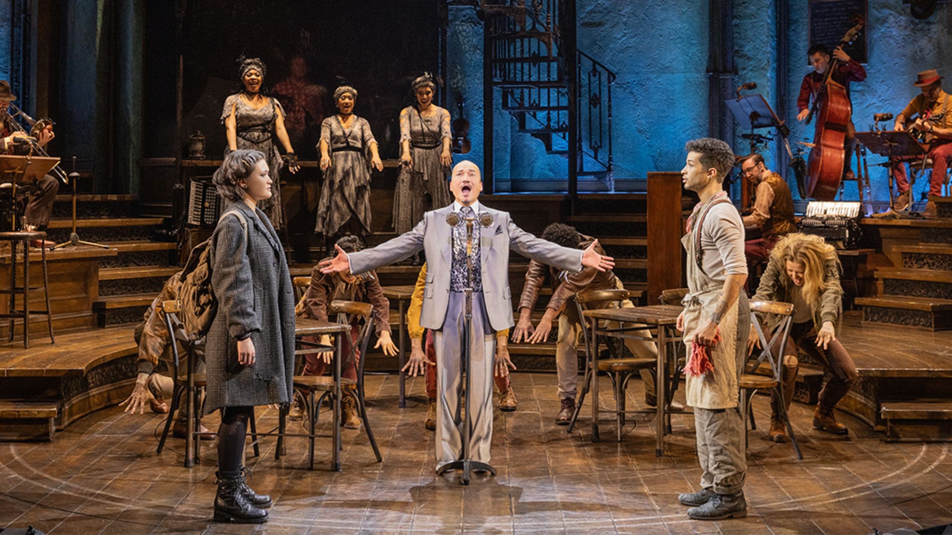 A man stands on stage, leading the cast of the musical, who are gathered around him in a vibrant display.