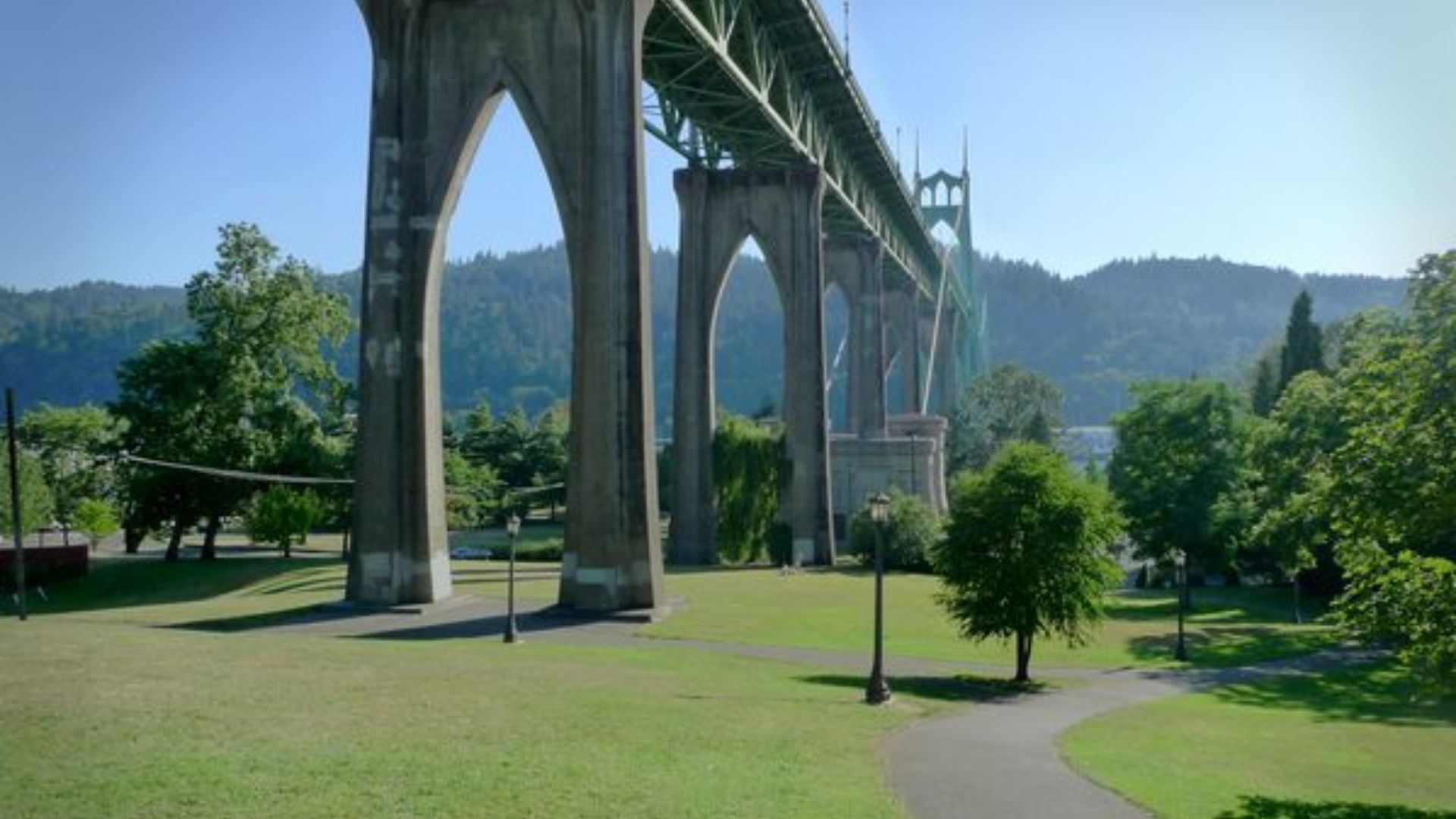 A bridge arches over a lush green park, surrounded by tall trees and open grassy areas.