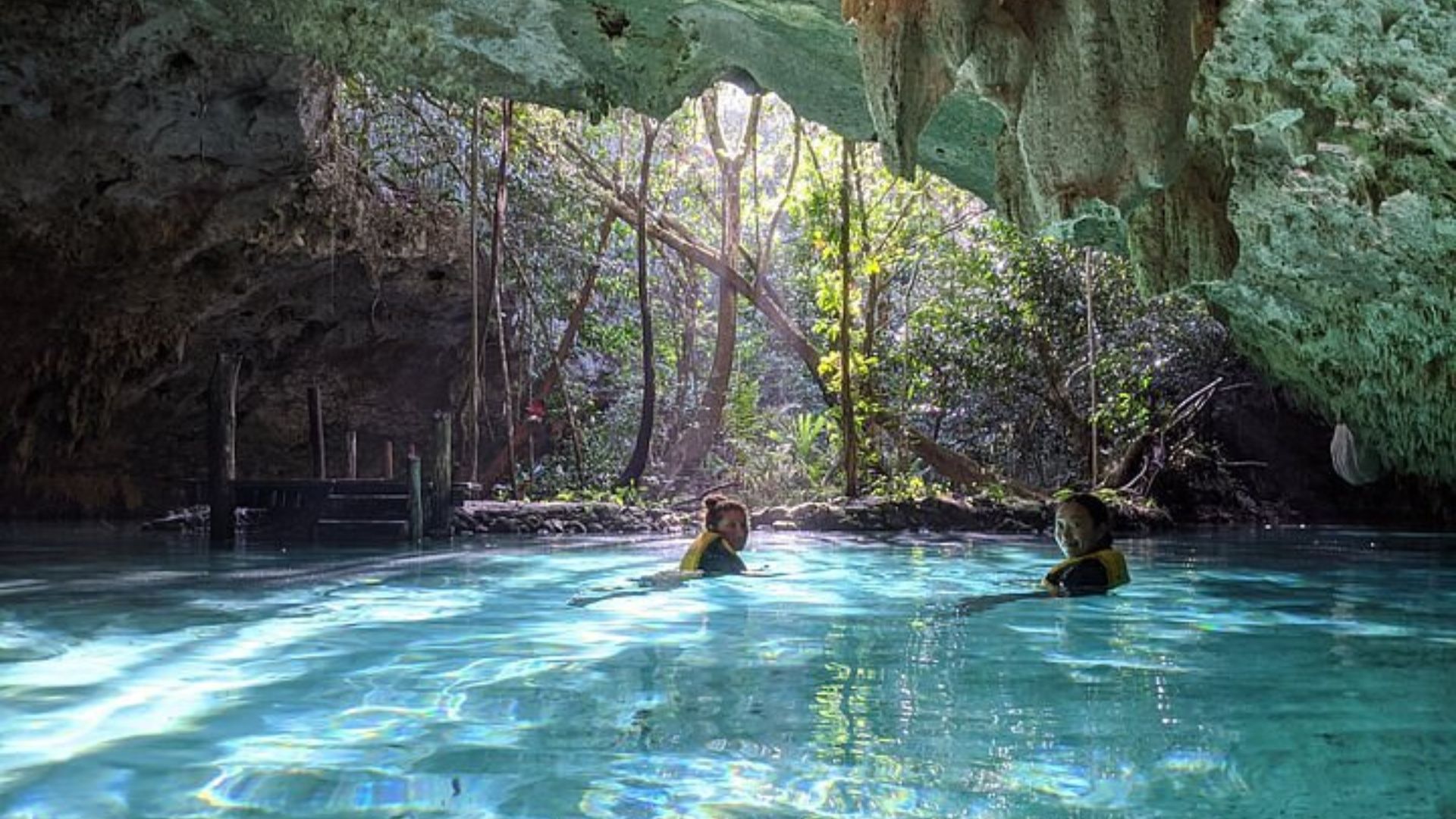 Two people swim in clear water inside a cave, surrounded by rocky walls and natural light filtering through the opening.