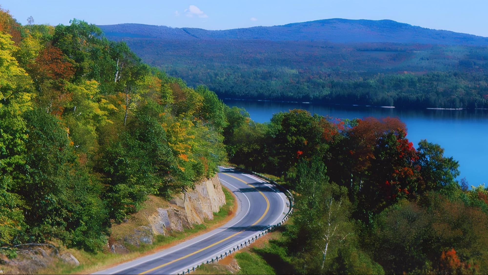 A winding road through mountains, surrounded by trees and a serene body of water, showcasing a picturesque landscape.