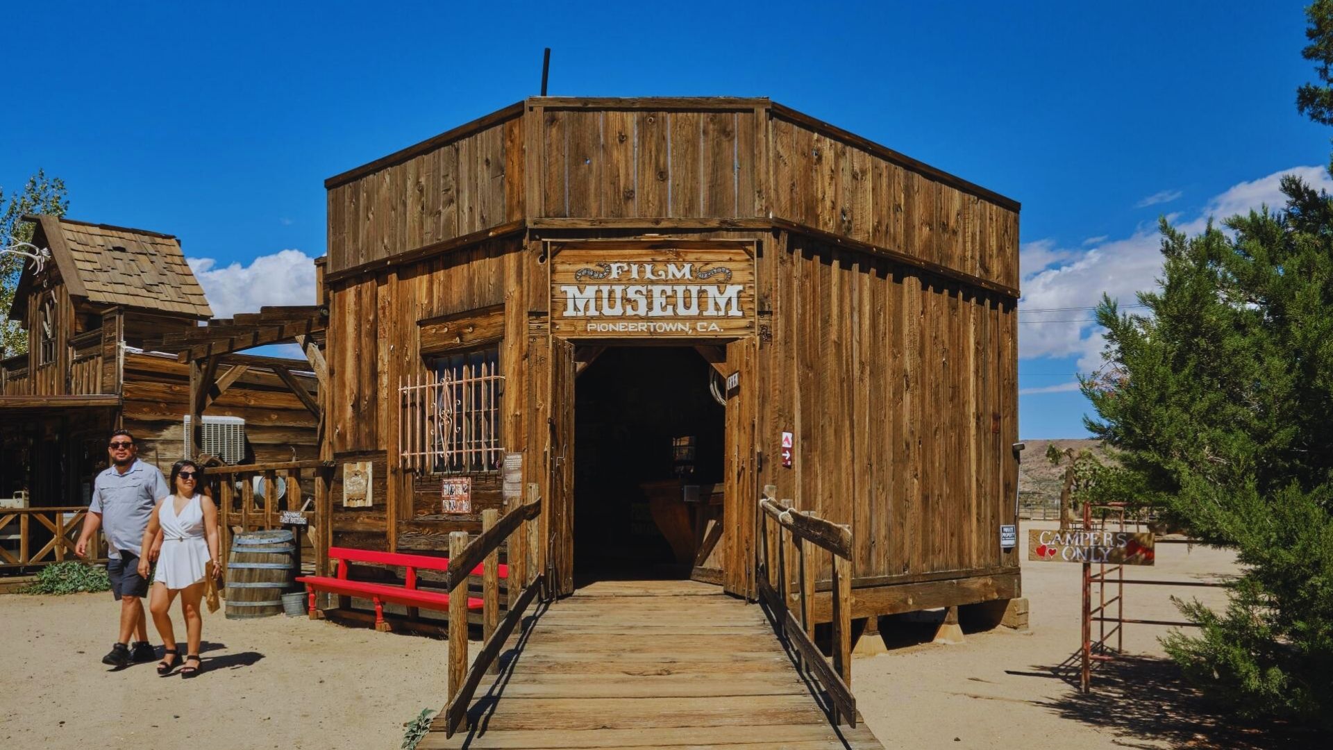 A couple approaches a rustic wooden building set against a desert landscape.