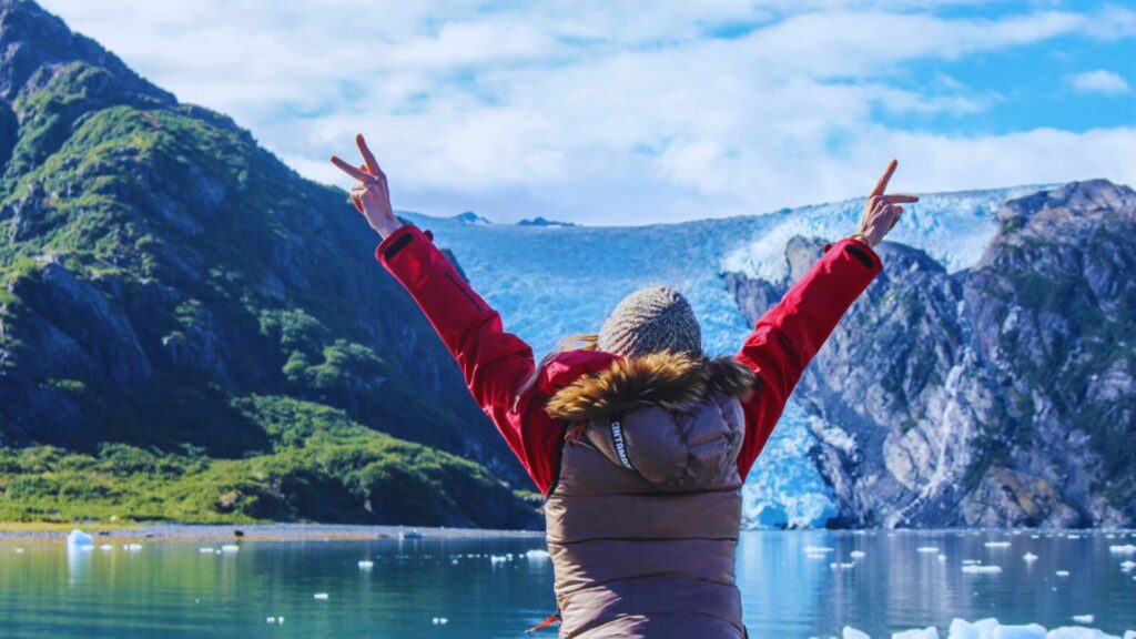 A woman stands with arms raised in front of a massive glacier, showcasing the grandeur of the icy landscape.