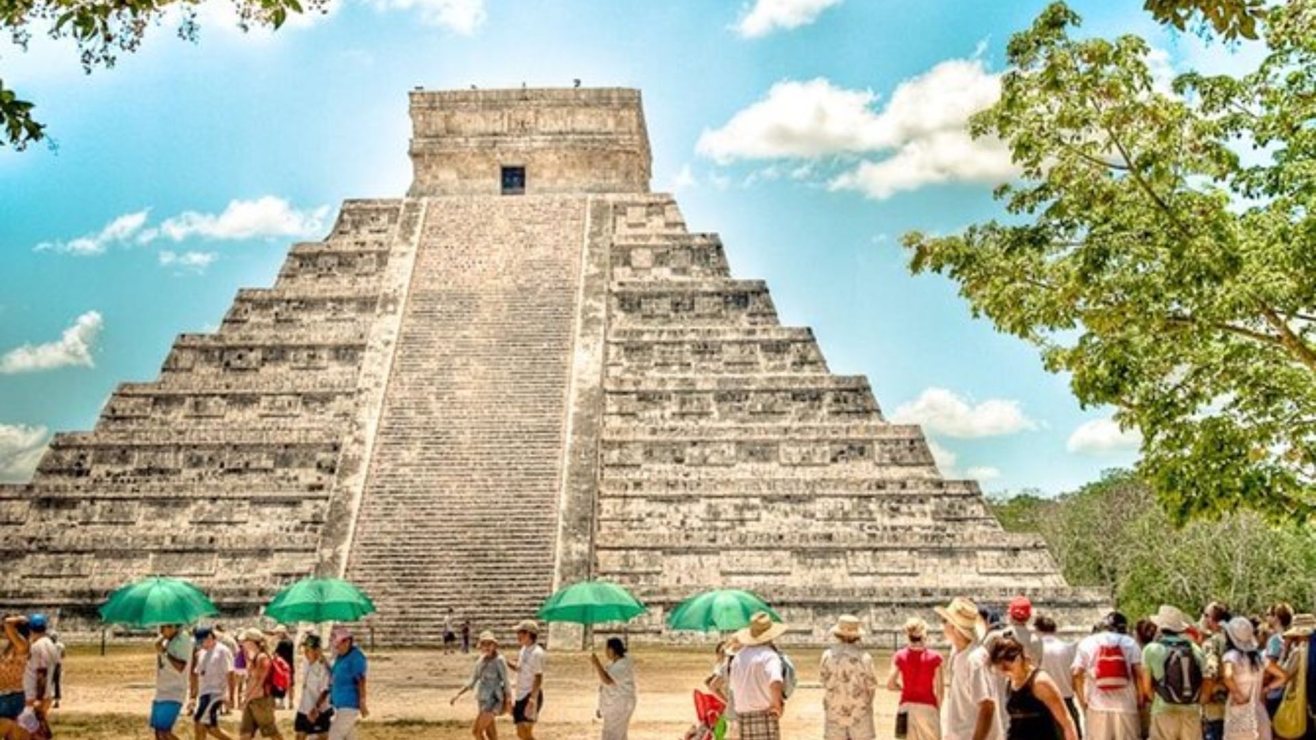 Tourists explore the ancient pyramid at Chichen Itza, surrounded by lush greenery and a clear blue sky.