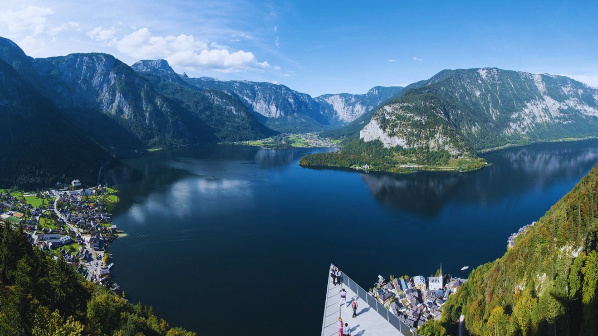 A panoramic view from the mountain summit, showcasing a serene lake surrounded by lush greenery and distant hills.