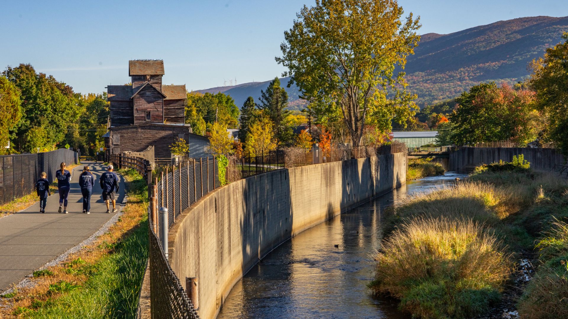 People stroll along a riverbank, with a bridge and clock tower visible in the background.