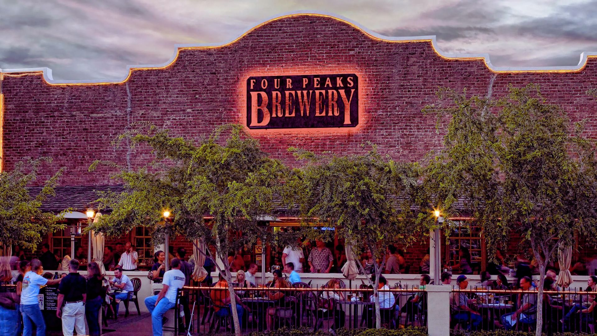 An American brewery at dusk, featuring glowing lights and people socializing on the patio under a twilight sky.