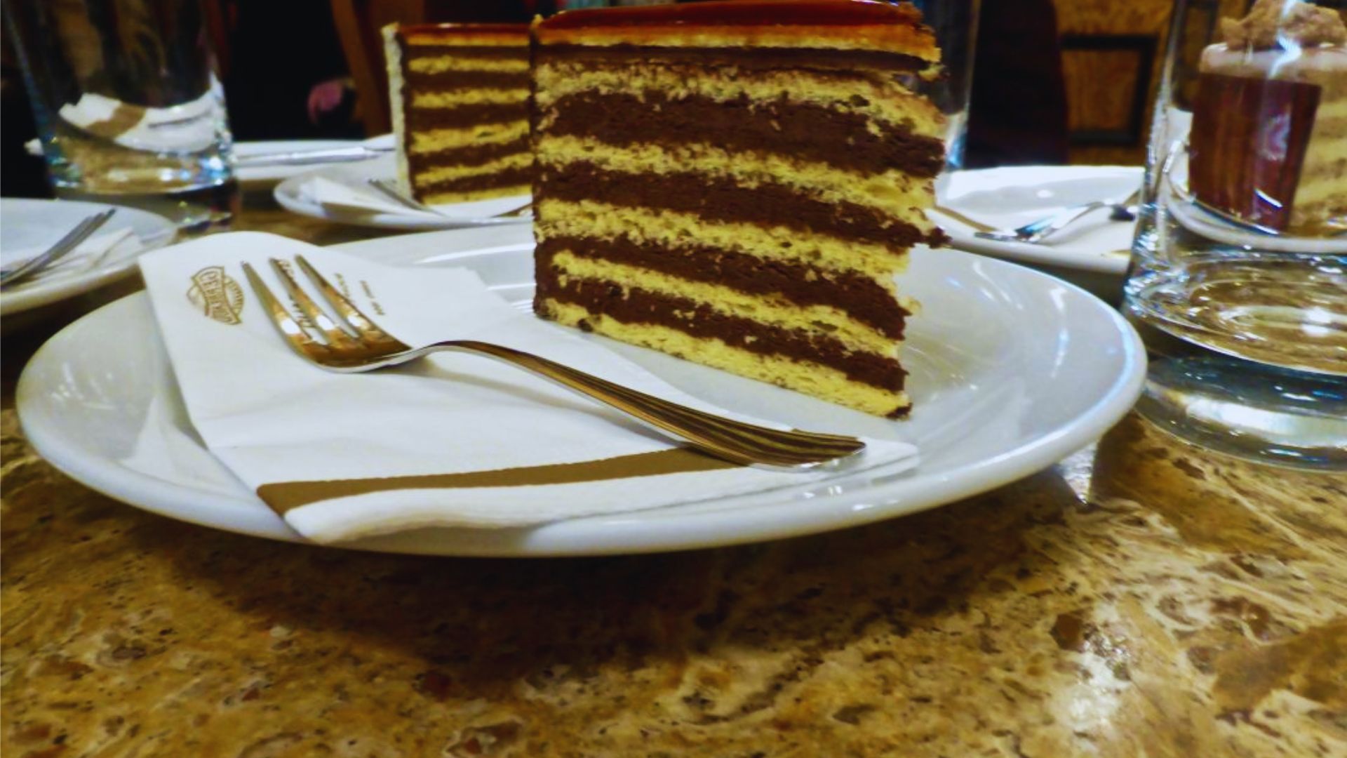 A slice of cake on a white plate, garnished with a cherry on top, set against a neutral background.