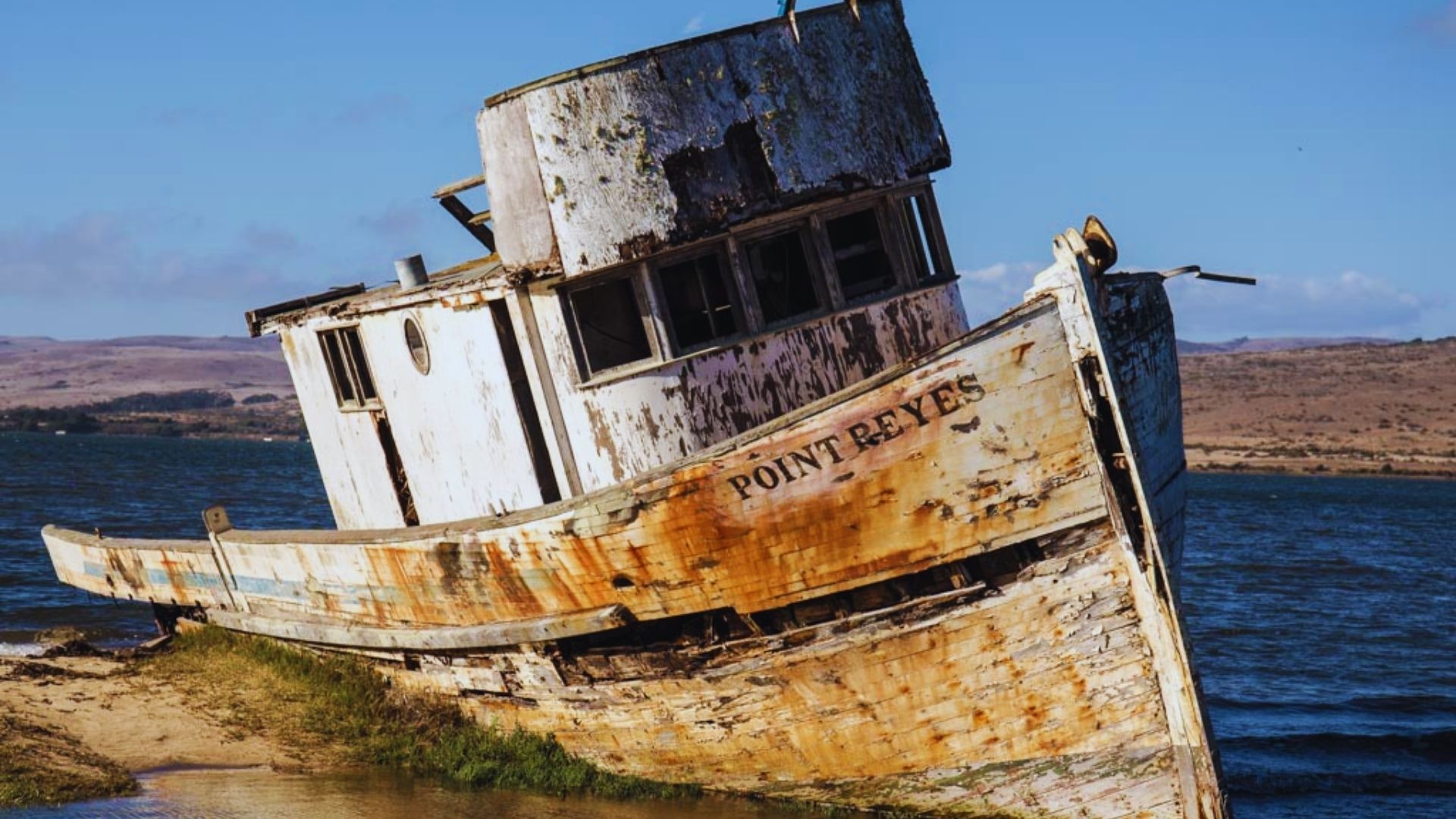 A weathered, rusty boat sits on the beach, partially buried in sand, with waves lapping nearby.