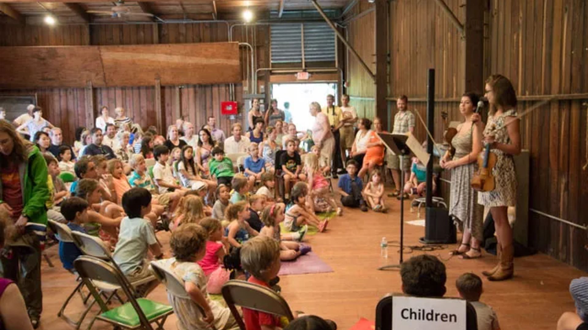 A group of children seated in chairs, attentively watching a performance on stage in front of them.