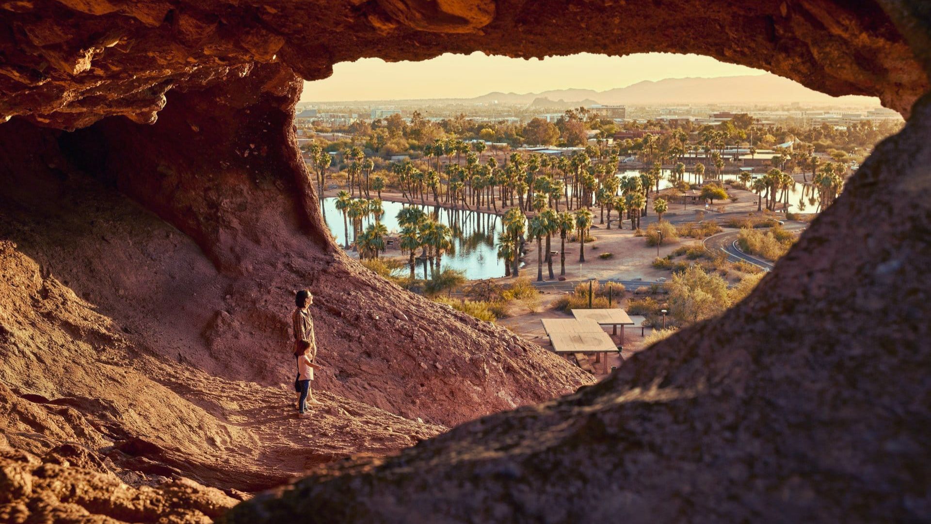 A person stands in a cave, gazing out at a sprawling cityscape illuminated by sunlight.