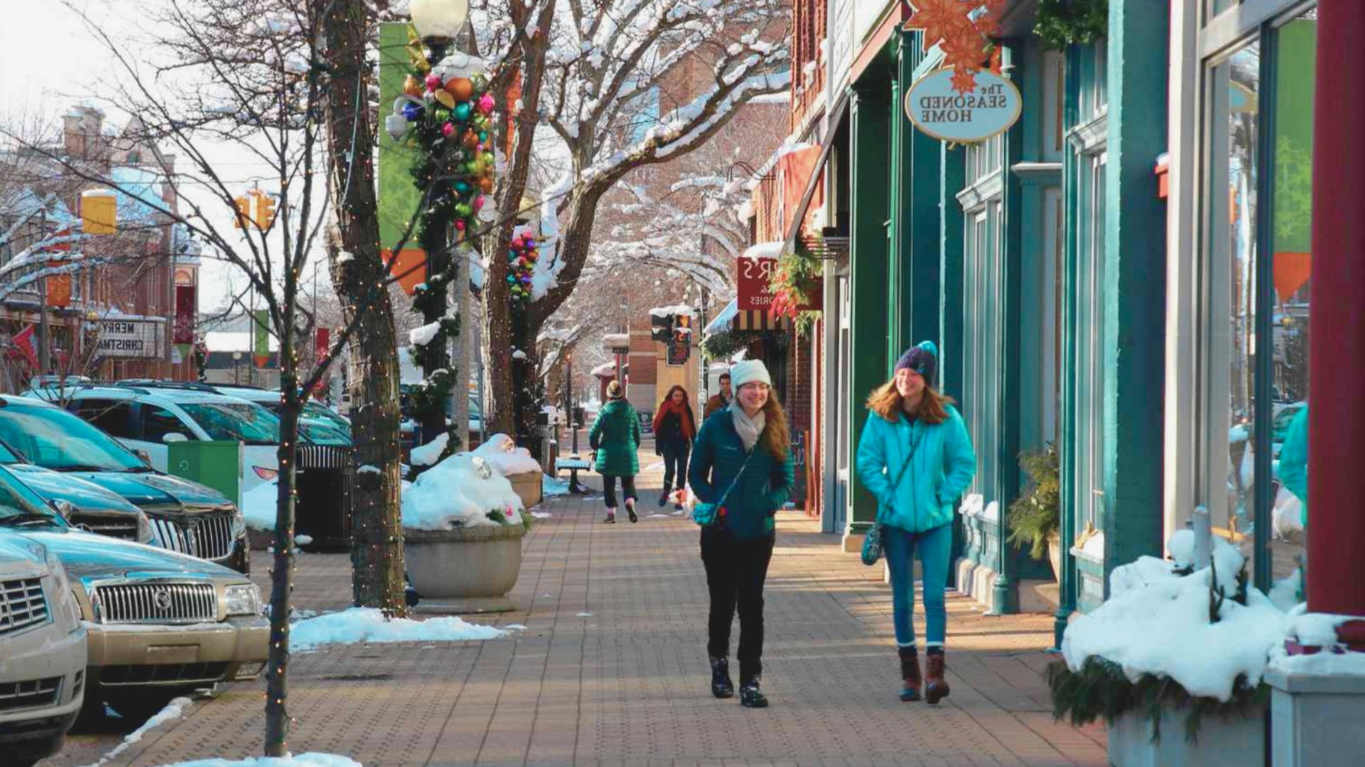 Two women walk side by side on a snowy sidewalk, bundled in winter coats and scarves.