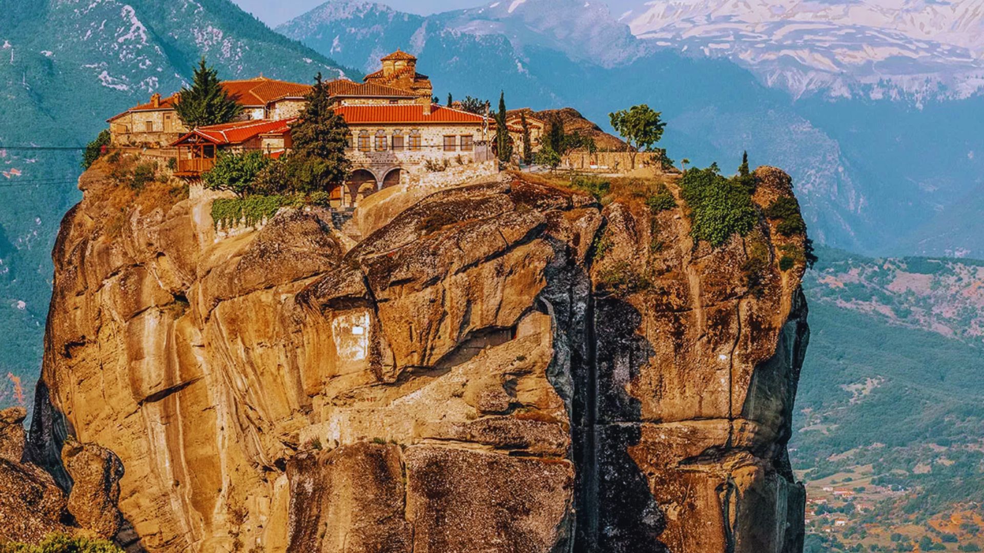A monastery perched atop a mountain in Greece, surrounded by scenic landscapes and clear blue skies.