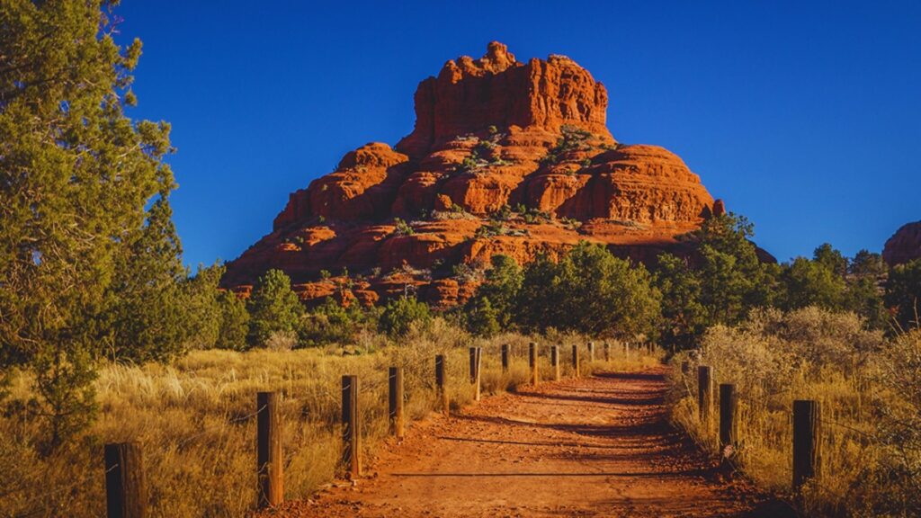 A dirt path stretches toward a prominent red rock formation, set against a backdrop of open sky.