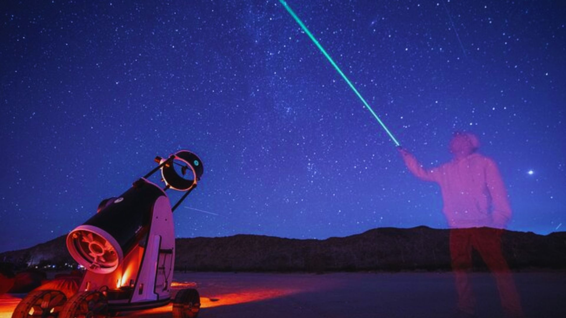 A man stands in front of a telescope, illuminated by a green light, gazing into the night sky.
