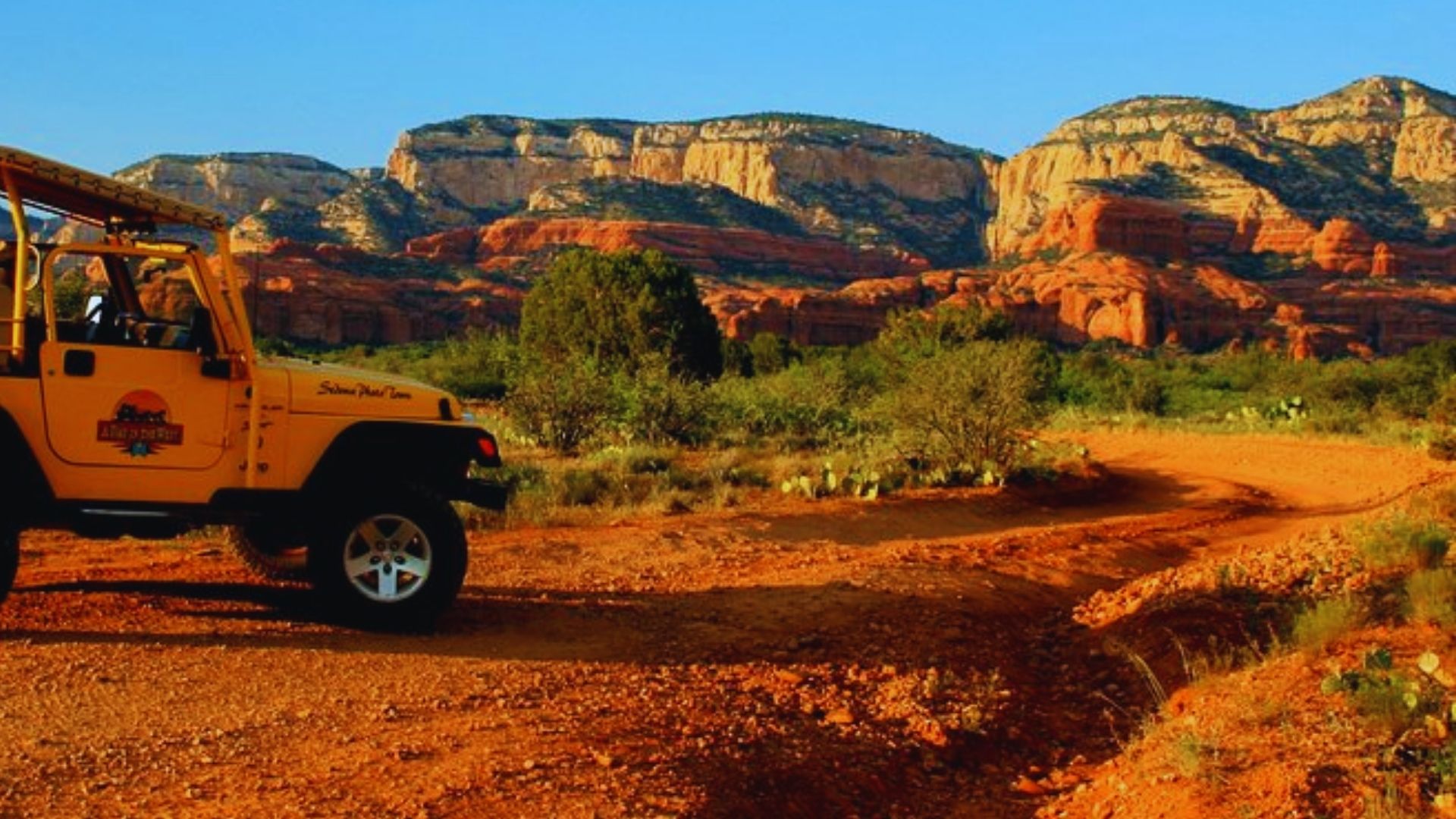A yellow jeep navigates a dirt road, framed by vibrant red rocks under a clear sky.