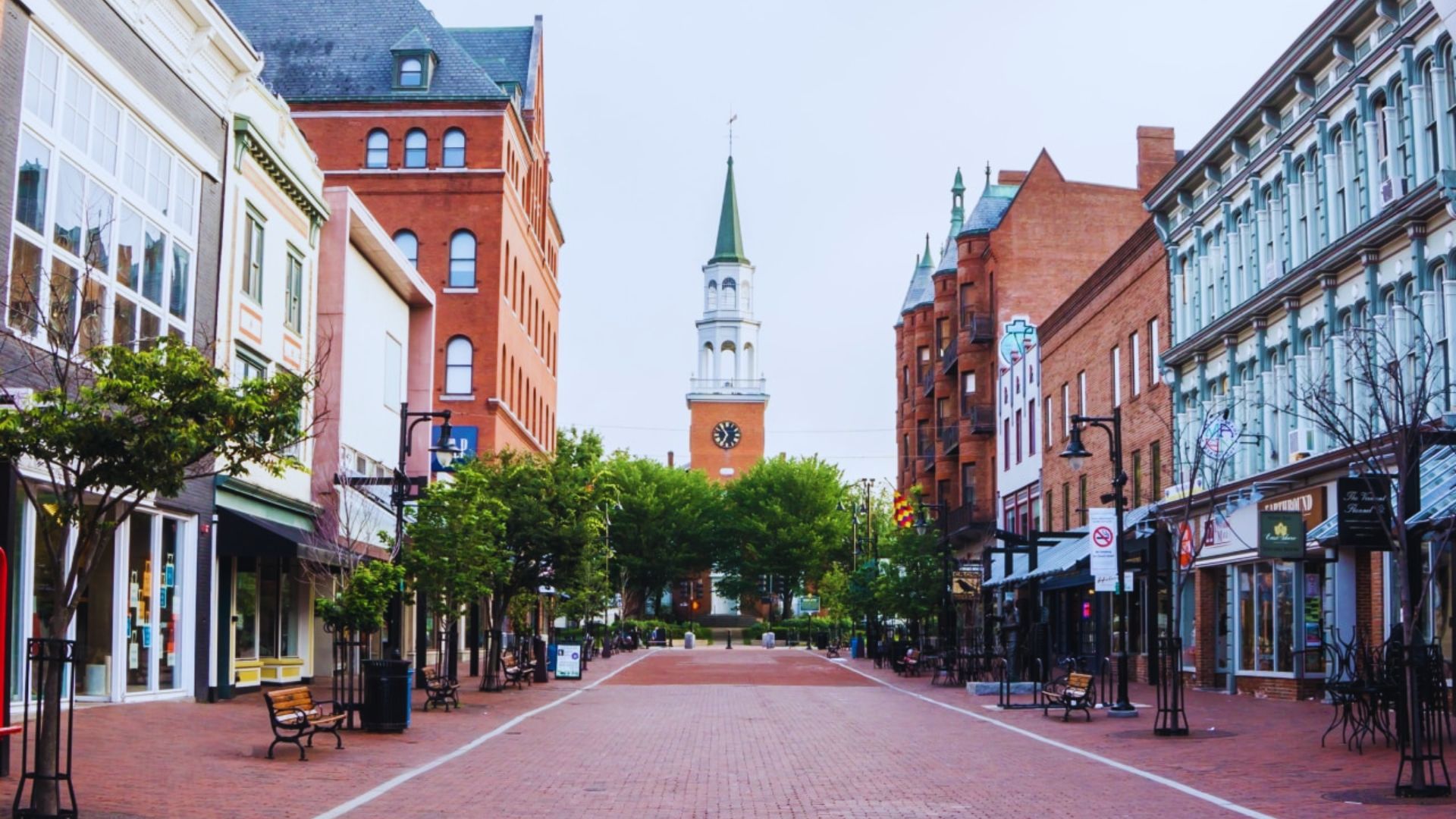 A city street lined with brick buildings, featuring a prominent steeple rising above the skyline.
