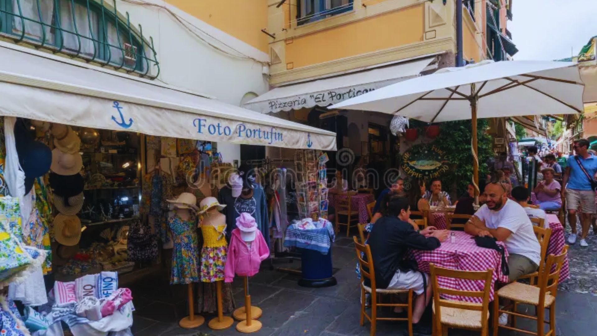 People seated at tables under umbrellas on a small street, enjoying their time in an outdoor café setting.
