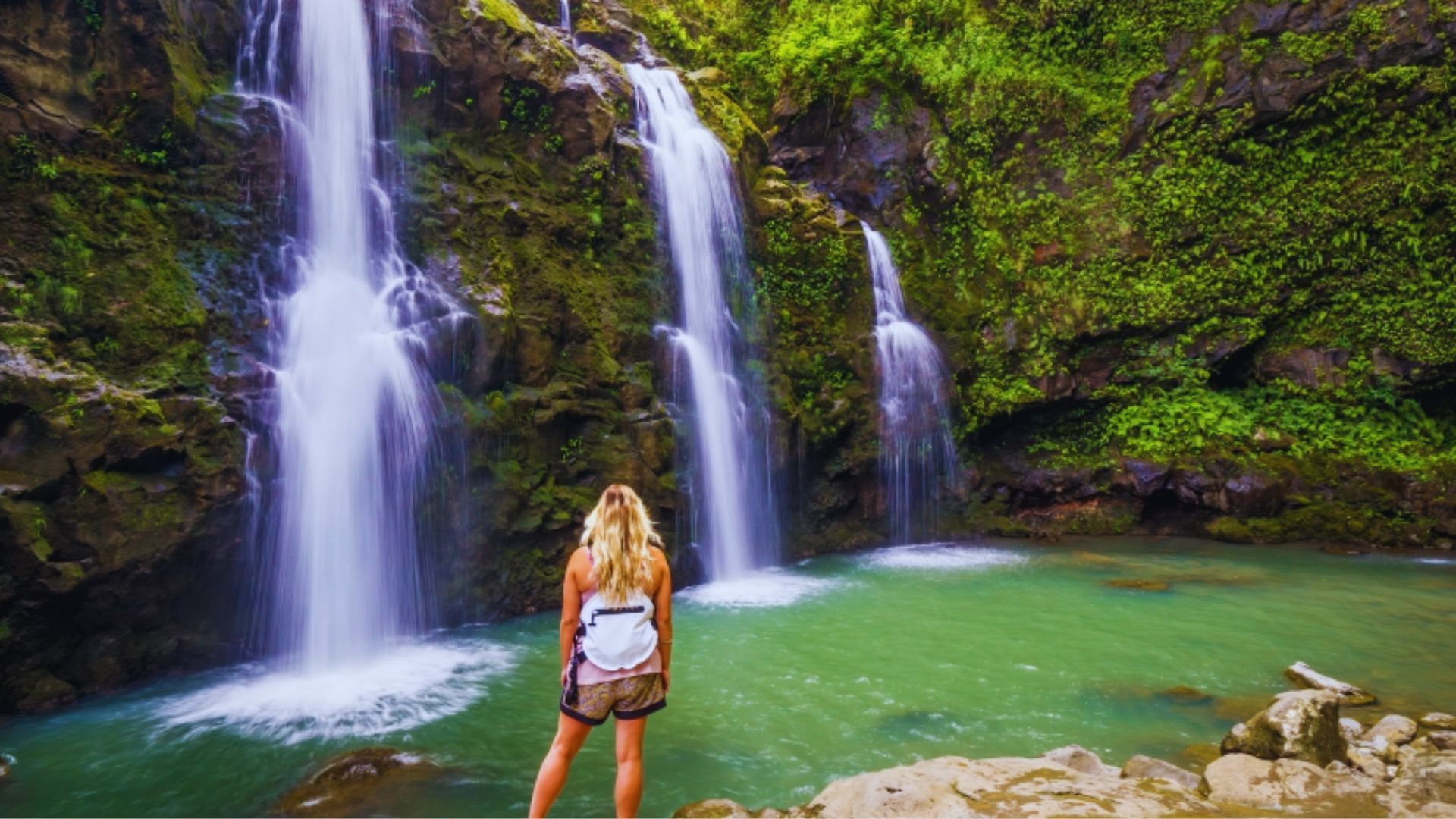 A woman stands in front of a lush jungle waterfall, surrounded by vibrant greenery and mist rising from the water.