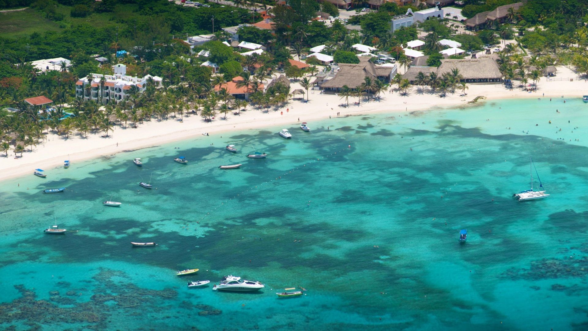 A scenic beach with boats anchored nearby and a resort visible in the distance under a clear blue sky.